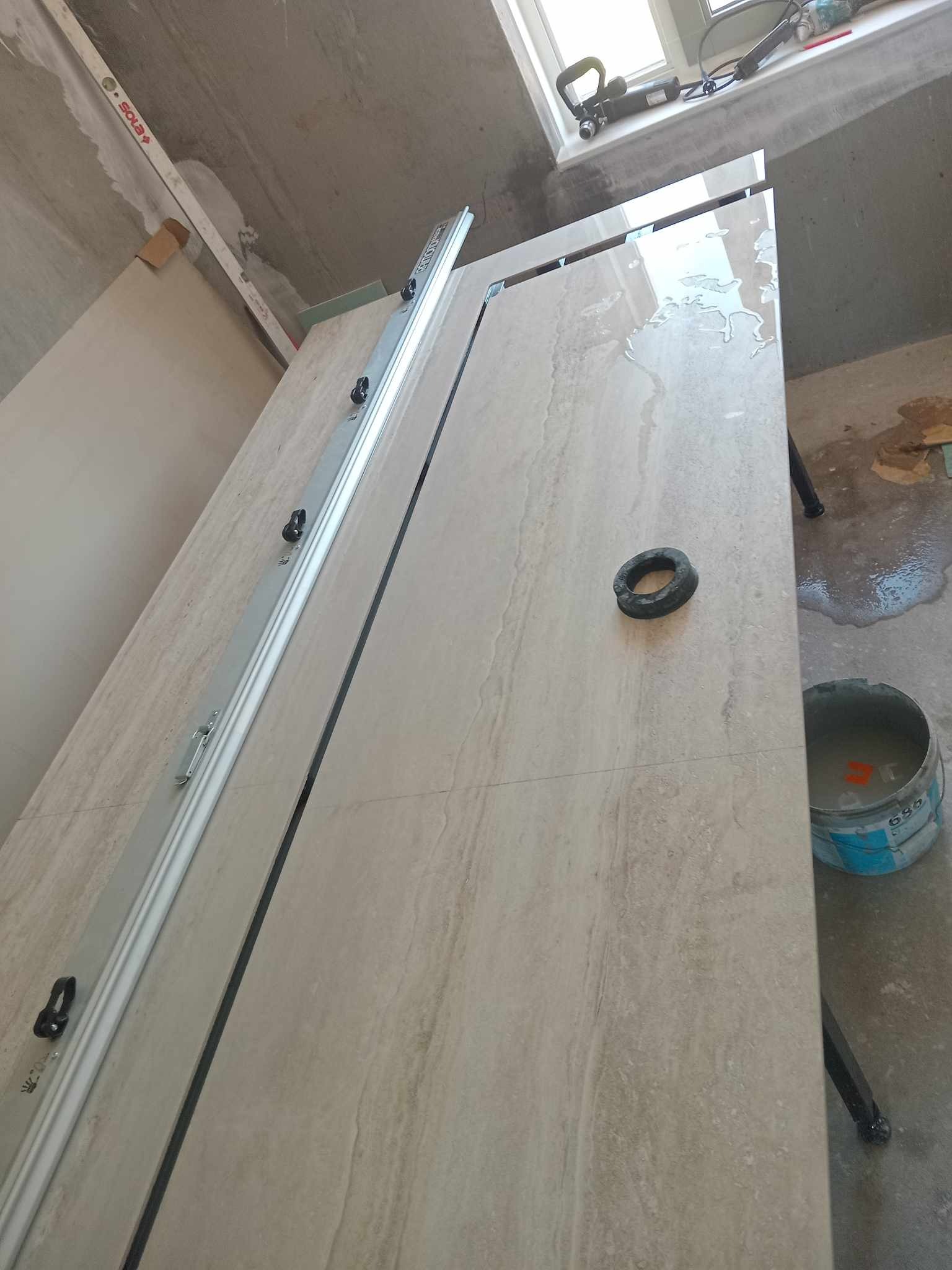 Układanie płytek podłogowych imitujących kamień, z użyciem poziomicy laserowej i wiadra z zaprawą, widoczne mokre powierzchnie i narzędzia na tle surowego wnętrza.