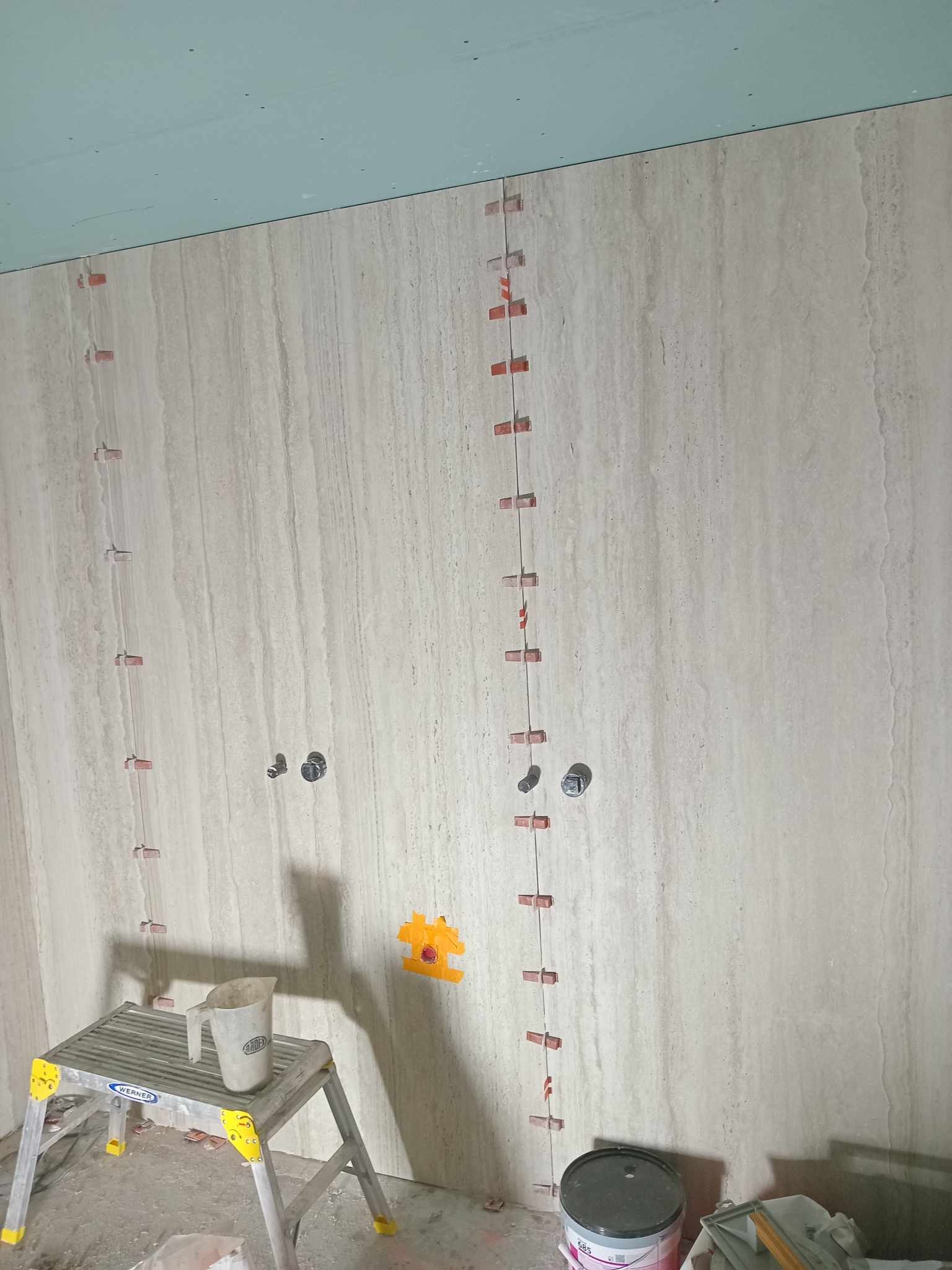 Ściana w trakcie układania płytek imitujących trawertyn, z widocznymi krzyżykami dystansowymi, instalacją wodną i poziomnicą z wiaderkiem na podeście.