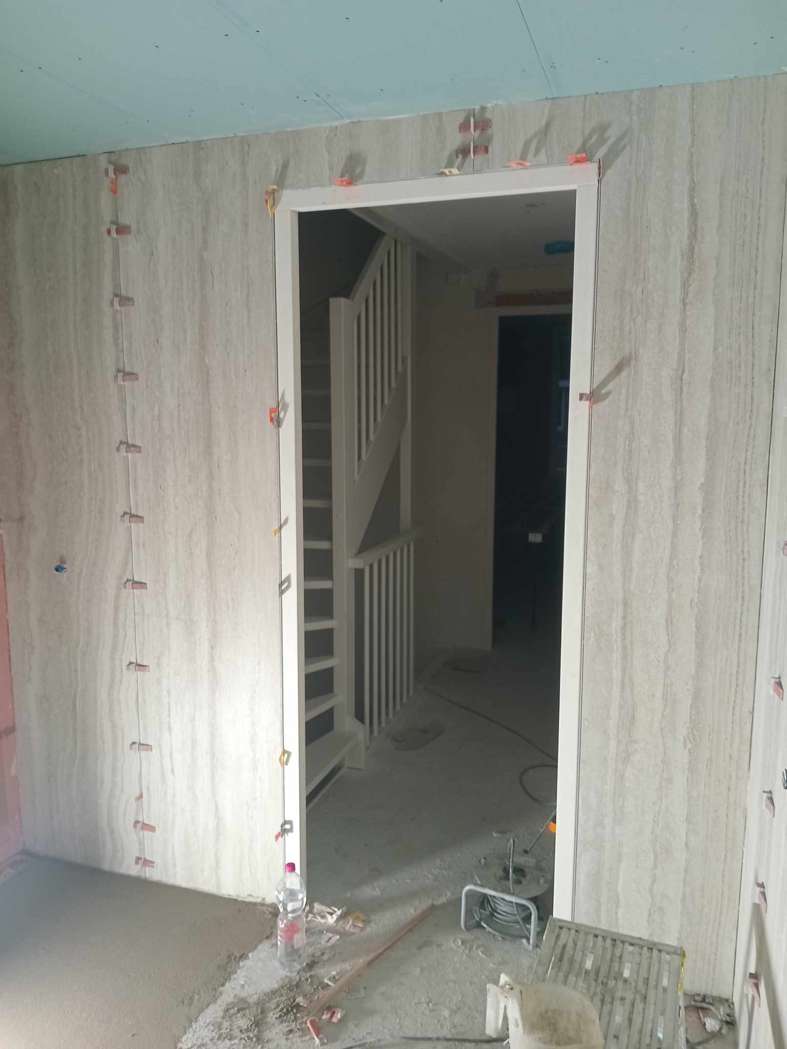 Montaż płytek imitujących trawertyn na ścianie, widoczne klipsy poziomujące, w tle schody i surowy stan wykończenia podłogi.