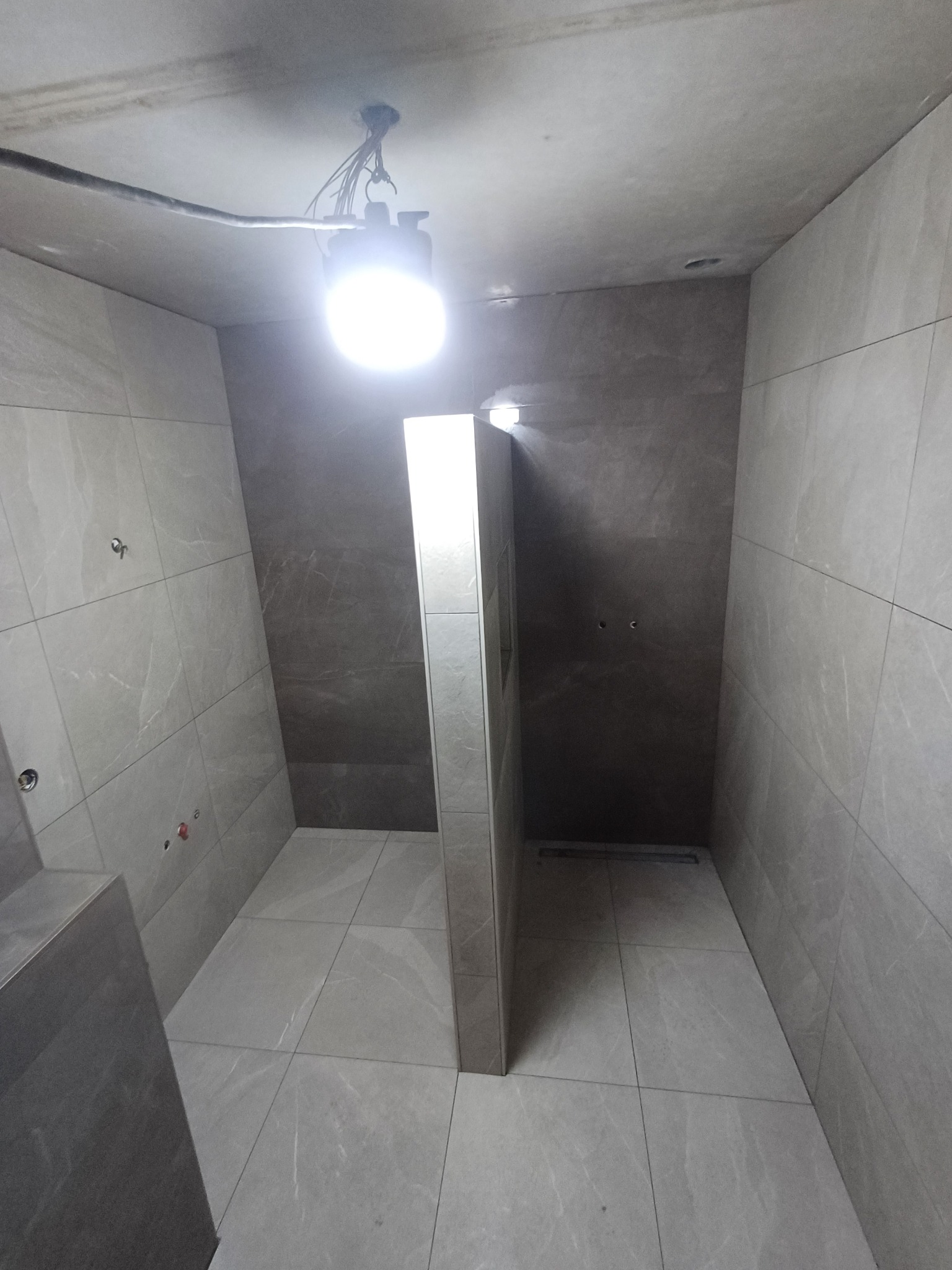 Wnętrze łazienki w trakcie wykańczania, widoczne szare płytki na ścianach i podłodze, zamontowany stelaż oddzielający prysznic, surowy sufit z wiszącym oświetleniem roboczym, brak armatury.