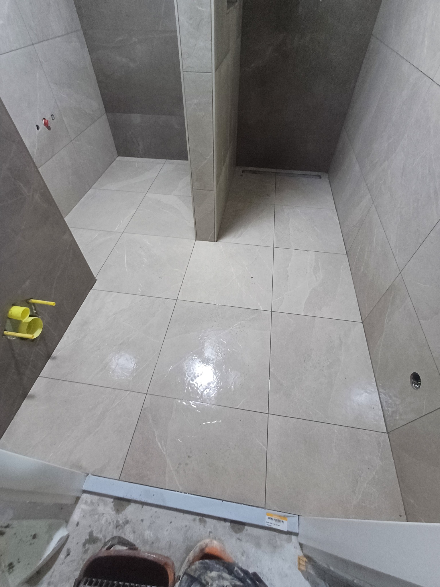 Wykończona kabina prysznicowa z szarymi płytkami imitującymi marmur, widoczne żółte rury instalacji wodnej i fragment butów roboczych w dolnej części kadru.