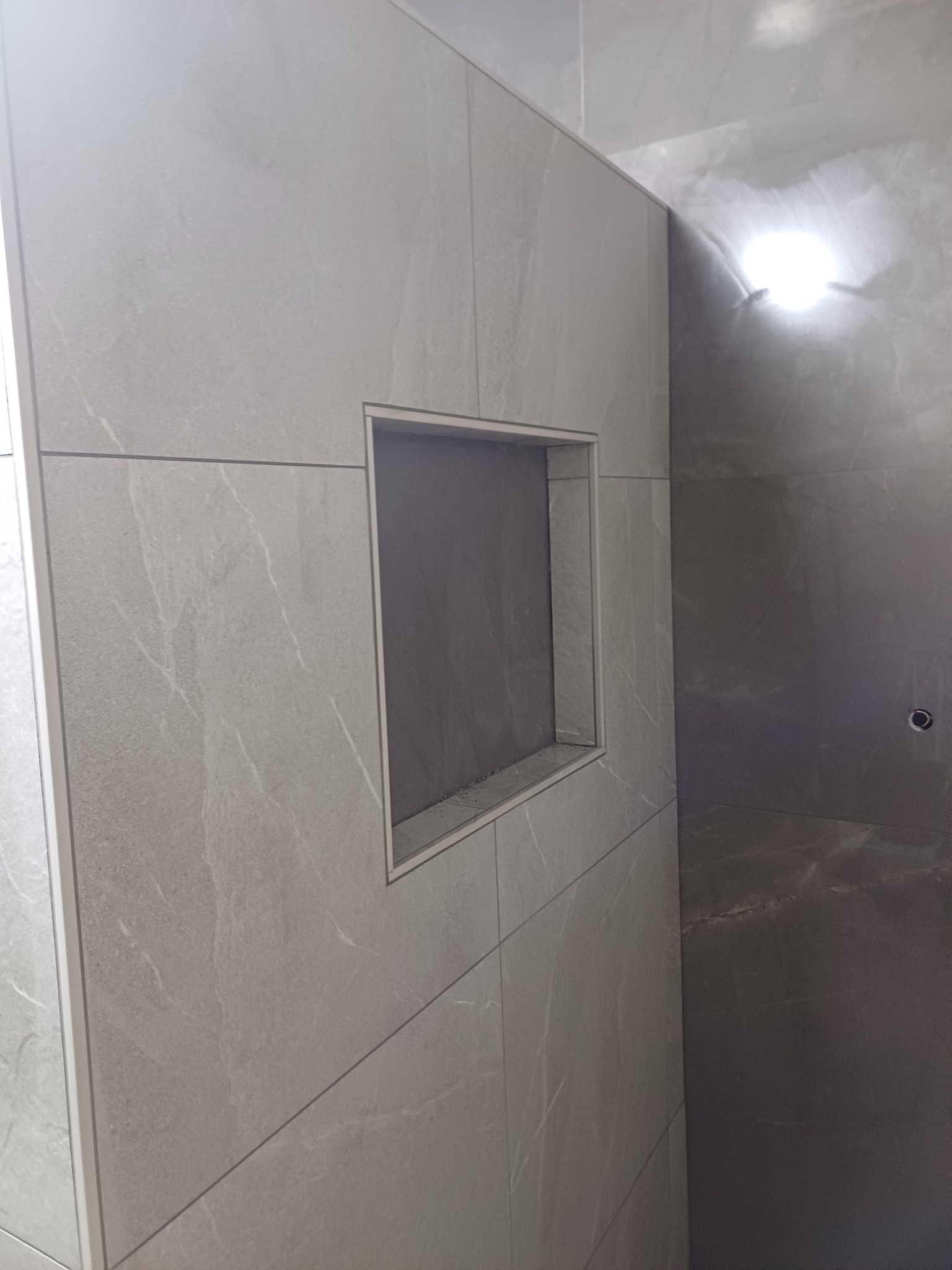 Ściana wyłożona szarymi płytkami w łazience w Warszkowie, z wnęką wykończoną tym samym materiałem, widoczne fugi i narożnik.