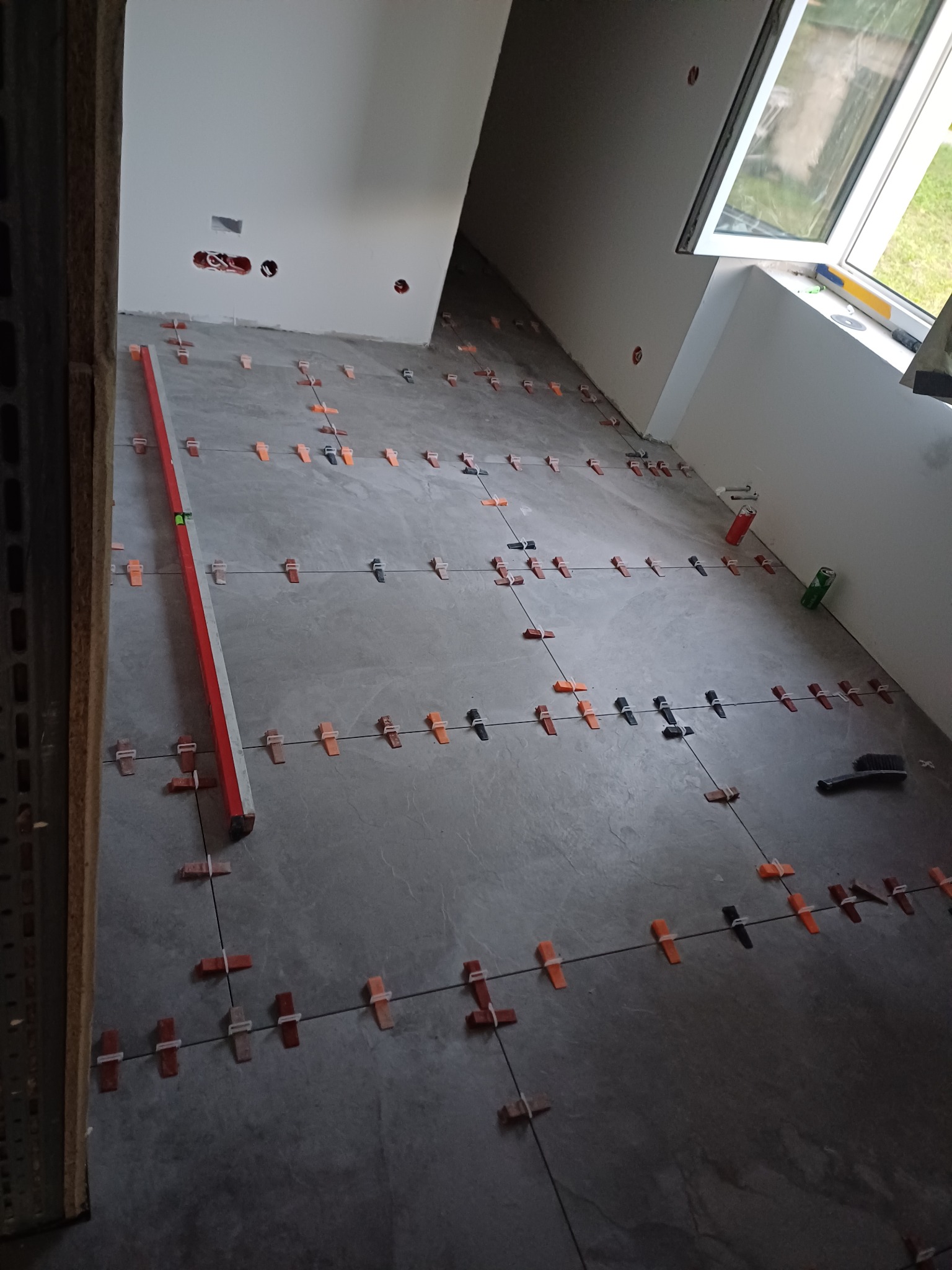 Szare płytki podłogowe w trakcie układania, z użyciem poziomicy i krzyżyków dystansowych, widok na surową ścianę z puszkami elektrycznymi i okno.