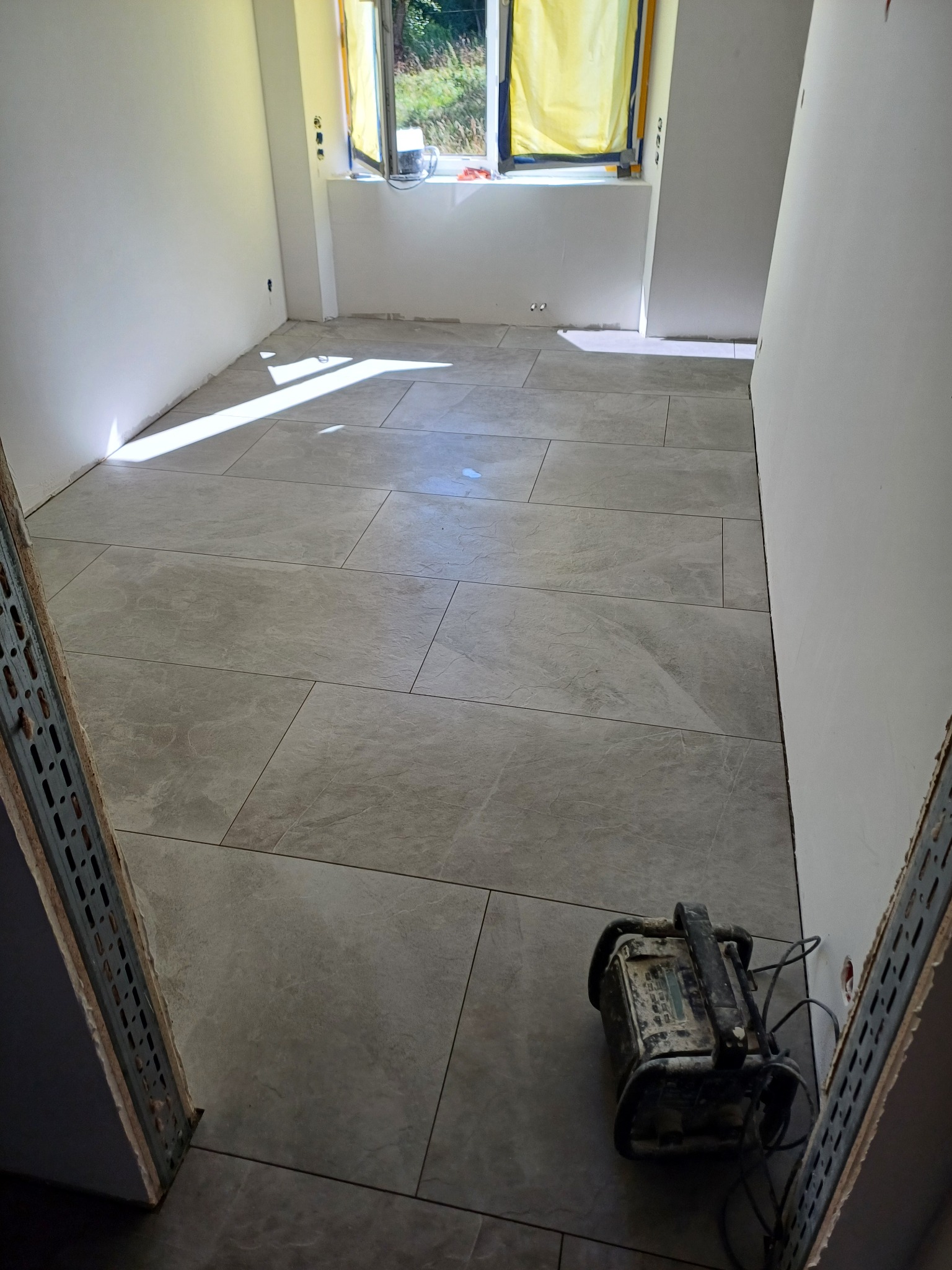 Ułożone szare płytki podłogowe w pomieszczeniu w trakcie remontu, widoczny brudny halogen budowlany i zabezpieczone okno folią.