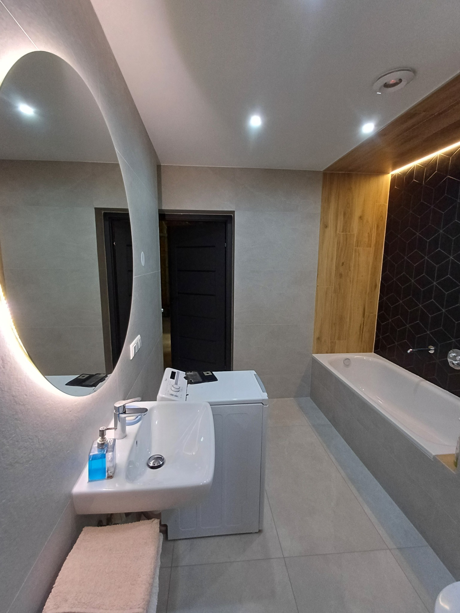 Nowoczesna łazienka z szarymi płytkami, owalnym lustrem z podświetleniem LED, pralką pod umywalką, drewnianą okładziną ściany i czarną mozaiką geometryczną nad wanną.