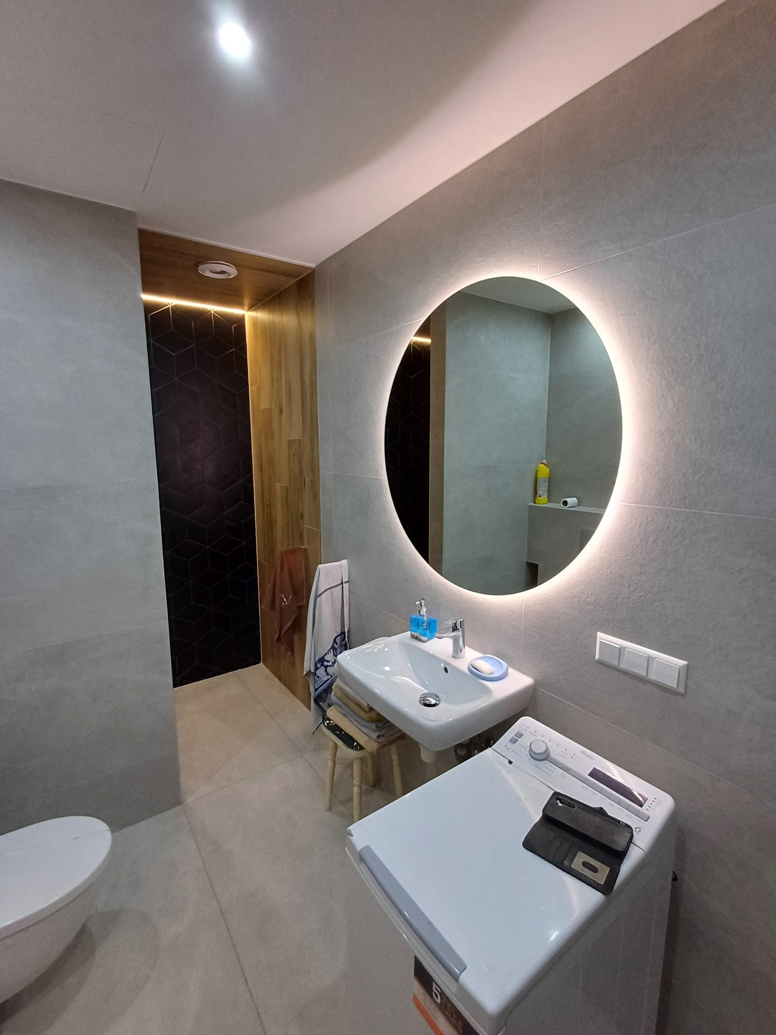 Nowoczesna łazienka z szarymi płytkami, okrągłym lustrem z podświetleniem LED, białą umywalką i pralką, akcentem z czarnych heksagonalnych płytek i drewnianymi panelami z oświetleniem.