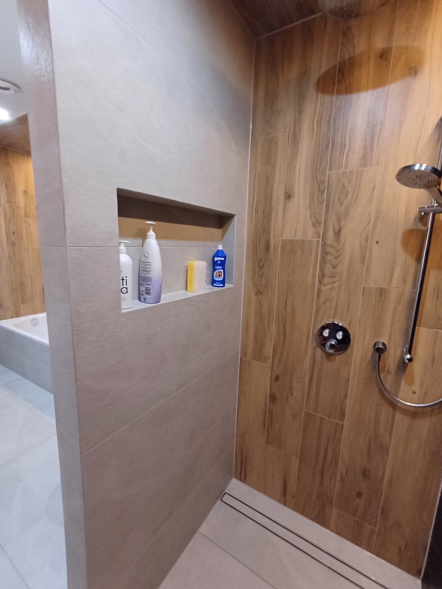 Nowoczesna łazienka z kabiną prysznicową typu walk-in, wykończona płytkami imitującymi drewno na ścianie z prysznicem i beżowymi płytkami na pozostałych ścianach, z wnęką w ścianie na kosmetyki...