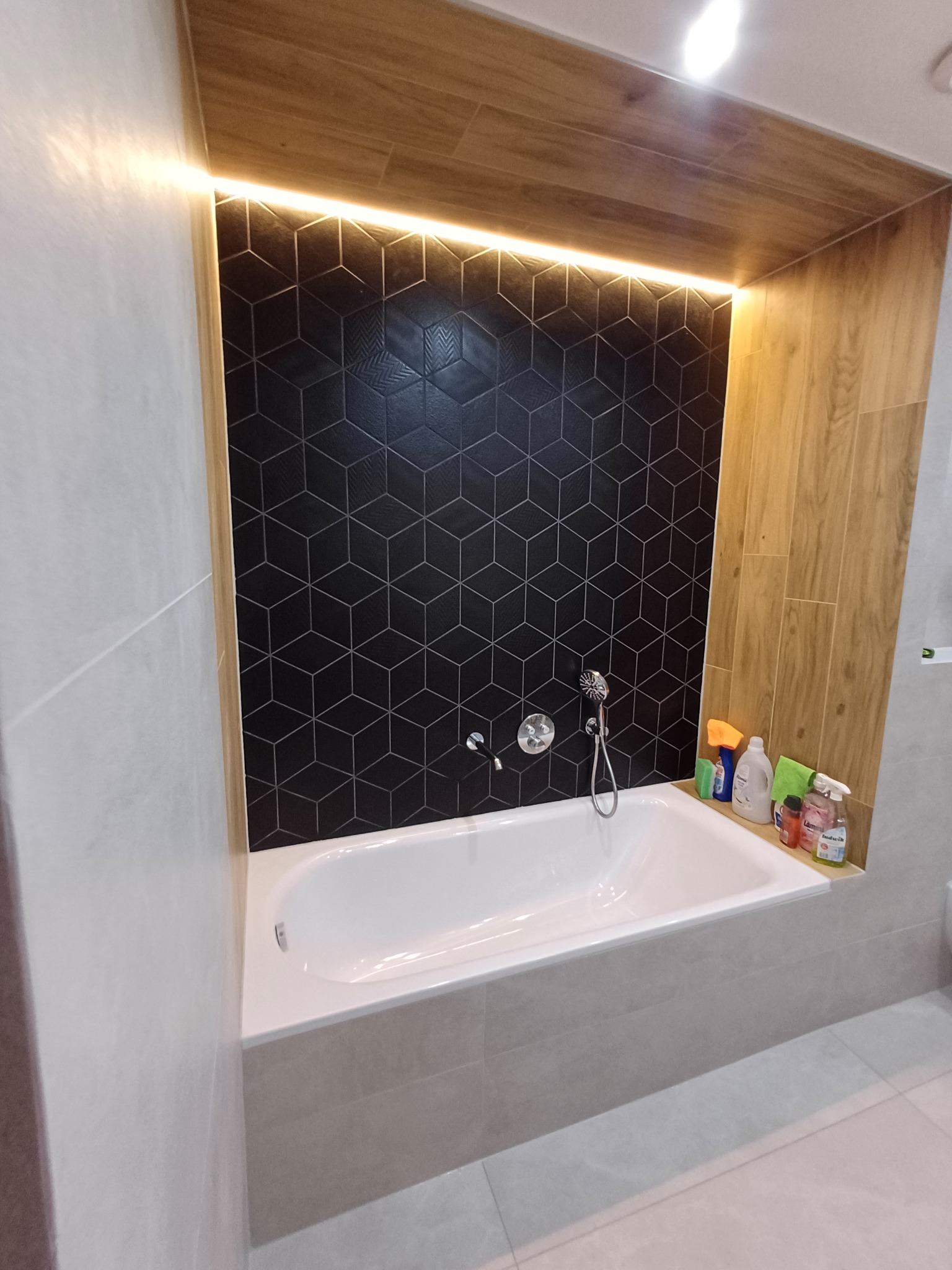 Nowoczesna łazienka z wanną obudowaną płytkami w odcieniach szarości, czarna ściana z geometrycznym wzorem płytek i drewnopodobna zabudowa z oświetleniem LED