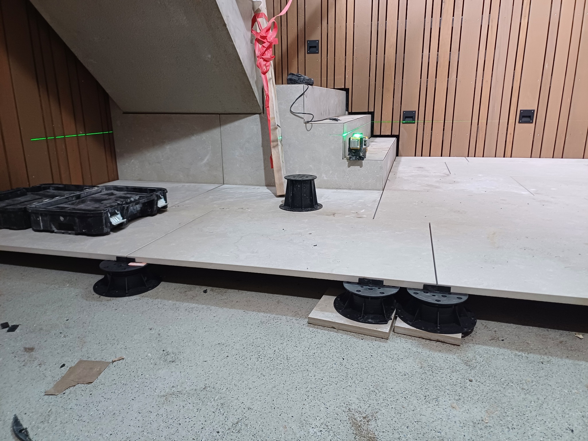 Układanie podłogi z dużych płyt na regulowanych wspornikach, widoczny laser poziomujący, schody w tle