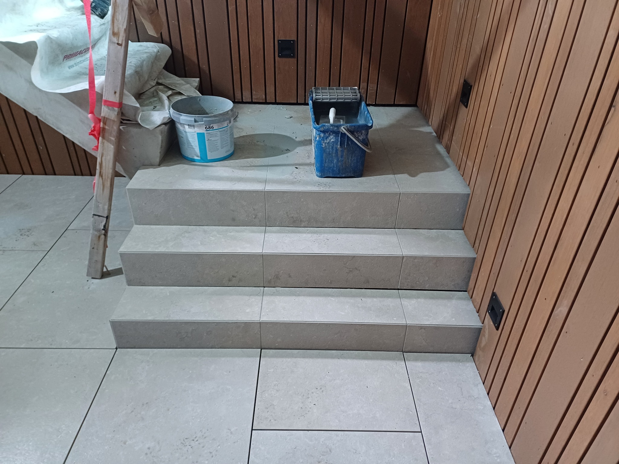 Wyłożone jasnymi płytkami schody prowadzące do drewnianej ściany, widoczny kubeł z narzędziami i wiadro z klejem do płytek.