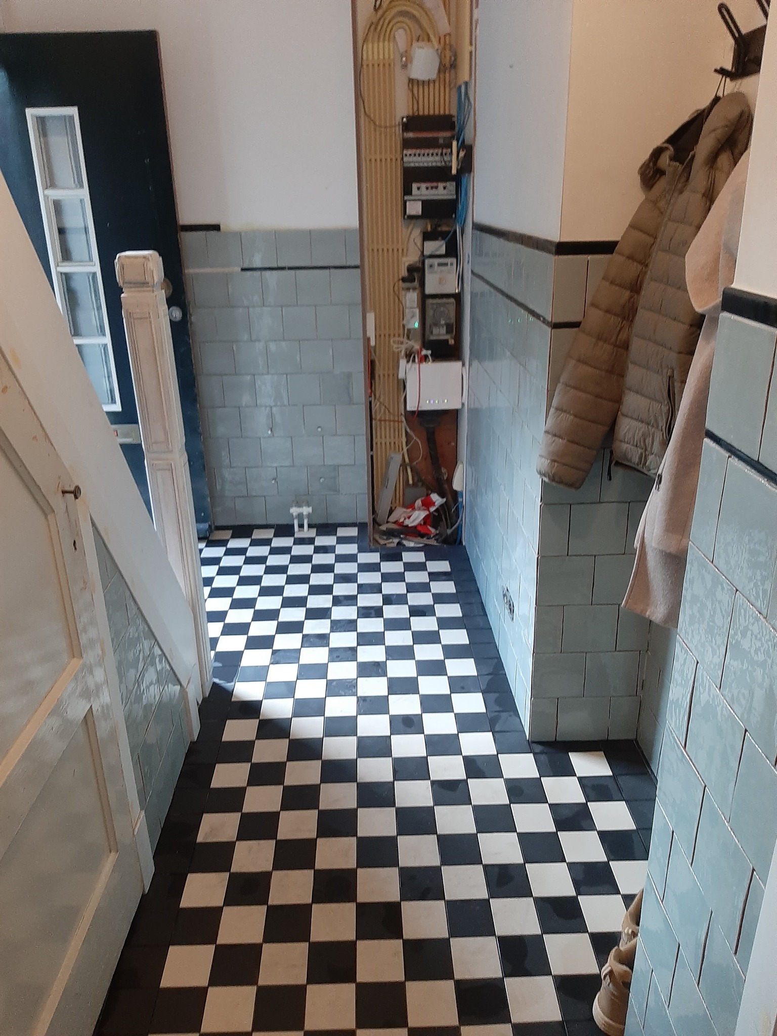 Korytarz z czarno-białą szachownicą na podłodze i ścianami wyłożonymi jasnoniebieskimi kafelkami, widoczne instalacje elektryczne na ścianie i kurtki wiszące na wieszaku.