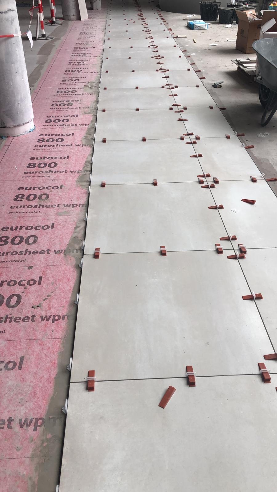 Układanie jasnych płytek podłogowych z użyciem systemu poziomowania, widoczna mata izolacyjna Eurocol 800 i narzędzia glazurnicze w tle.