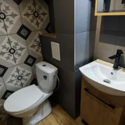 Nowoczesna łazienka po remoncie z płytkami w geometryczne wzory, drewnopodobną podłogą, białą umywalką z szafką i toaletą.