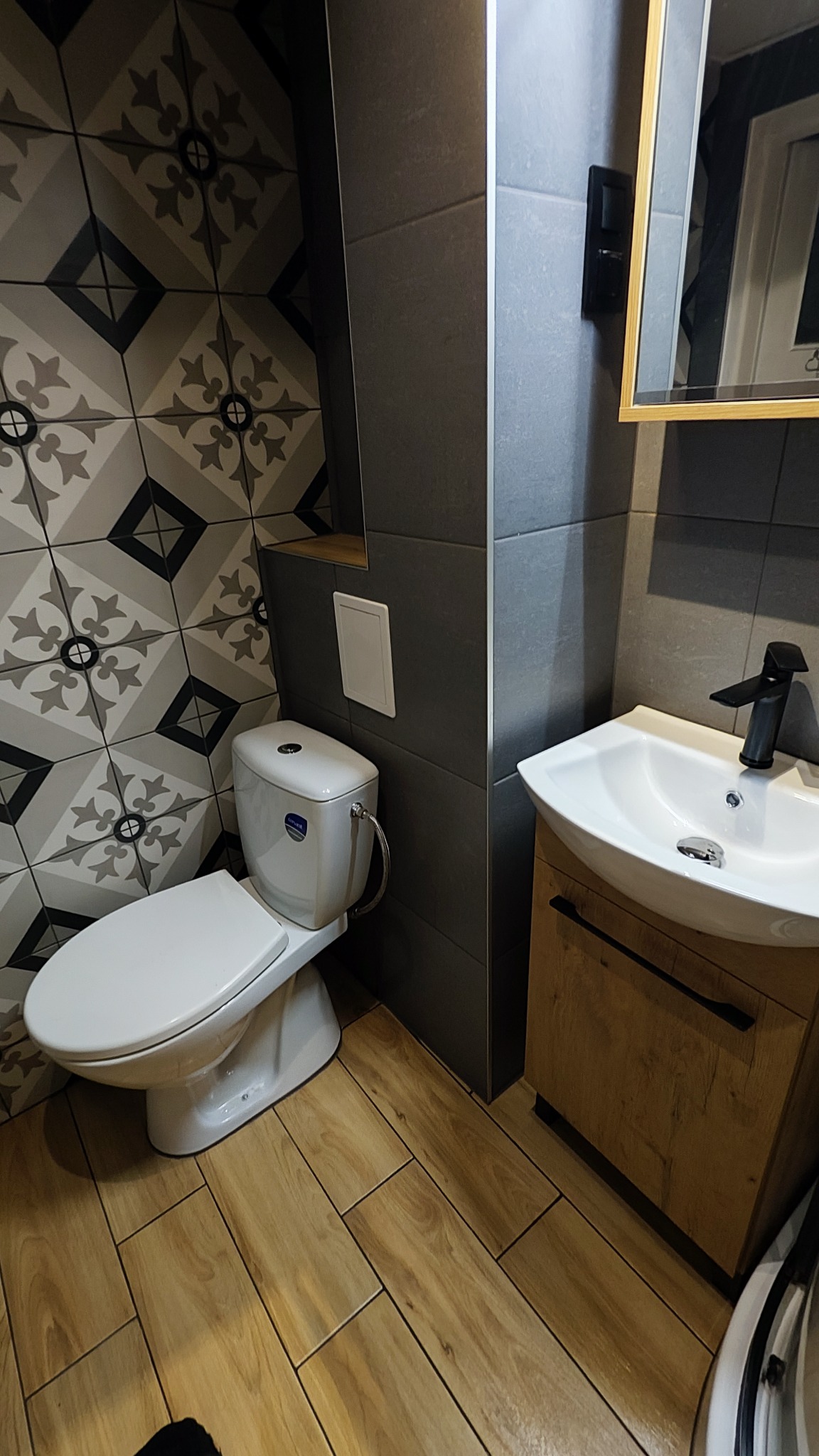 Nowoczesna łazienka po remoncie z płytkami w geometryczne wzory, drewnopodobną podłogą, białą umywalką z szafką i toaletą.
