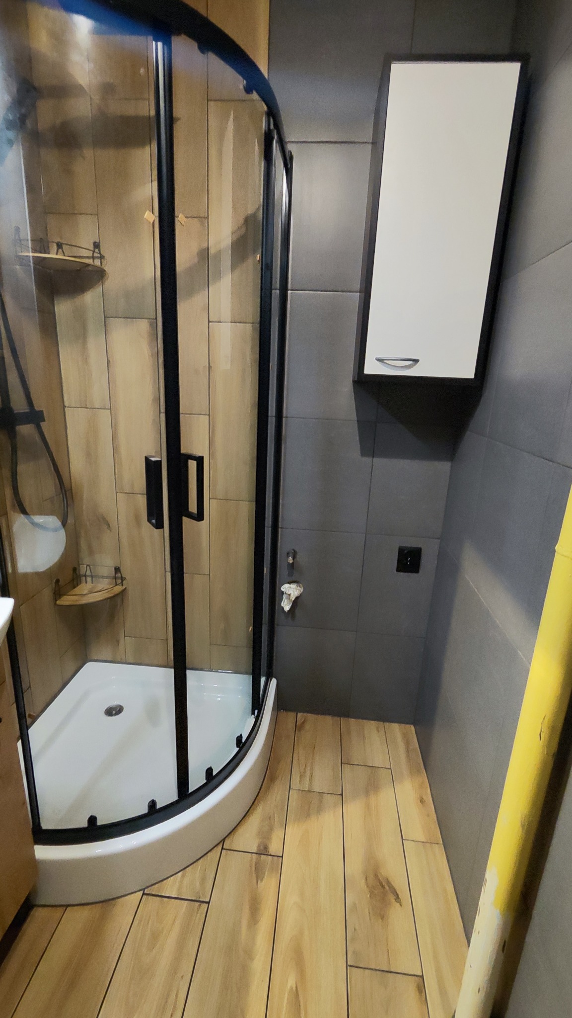 Wykończona kabina prysznicowa z czarnymi profilami w łazience w Gnieźnie, z drewnopodobnymi płytkami na ścianach i podłodze oraz szafką wiszącą nad podłogą.