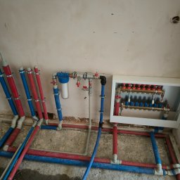 Kamil Rynkiewicz - Instalacja ogrzewania podłogowego z rozdzielaczem w białej obudowie i filtrem wody, rury czerwone i niebieskie, surowy stan wykończenia ścian i podłogi.