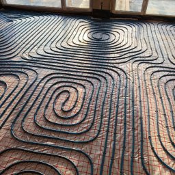 Kamil Rynkiewicz - Niebieskie rury ogrzewania podłogowego ułożone spiralnie na folii z czerwoną siatką, widoczne okno i fragment surowego betonu. Oświetlenie naturalne, ciepłe.