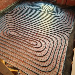 Kamil Rynkiewicz - Niebieskie rury ogrzewania podłogowego ułożone spiralnie na folii z czerwoną siatką w surowym wnętrzu. Widoczne fragmenty ceglanej ściany i okno.