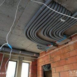 Kamil Rynkiewicz - Szereg szarych, karbowanych rur instalacyjnych przymocowanych do sufitu w surowym stanie, z widoczną czerwoną cegłą i oknem. Instalacja w trakcie realizacji.