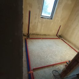 Kamil Rynkiewicz - Wnętrze w trakcie prac hydraulicznych: czerwono-niebieskie rury ułożone wzdłuż ścian i pionowo, surowy beton, okno dachowe. Widok z góry, etap instalacji.