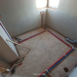 Kamil Rynkiewicz - Instalacja wodna i kanalizacyjna w surowym pomieszczeniu. Rury PEX w kolorze niebieskim i czerwonym ułożone wzdłuż ścian i pod oknem. Widoczne narzędzia hydrauliczne.