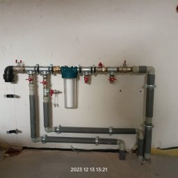Kamil Rynkiewicz - Szara instalacja wodna z filtrem i zaworami na białej ścianie. Rury mocowane obejmami. Widoczne przyłącza i elementy instalacji. Surowy stan wykończenia wnętrza.