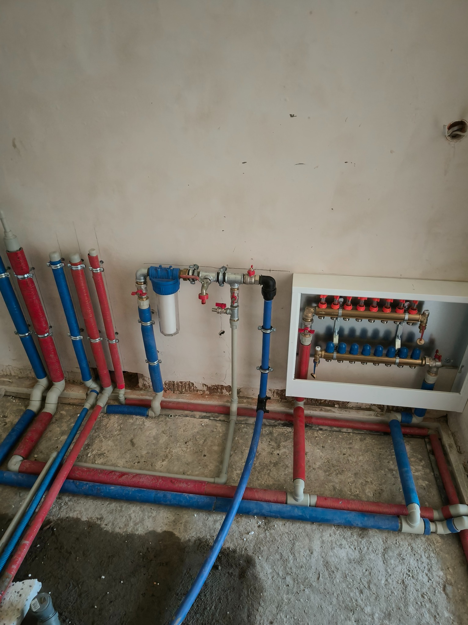 Instalacja ogrzewania podłogowego z rozdzielaczem w białej obudowie i filtrem wody, rury czerwone i niebieskie, surowy stan wykończenia ścian i podłogi.