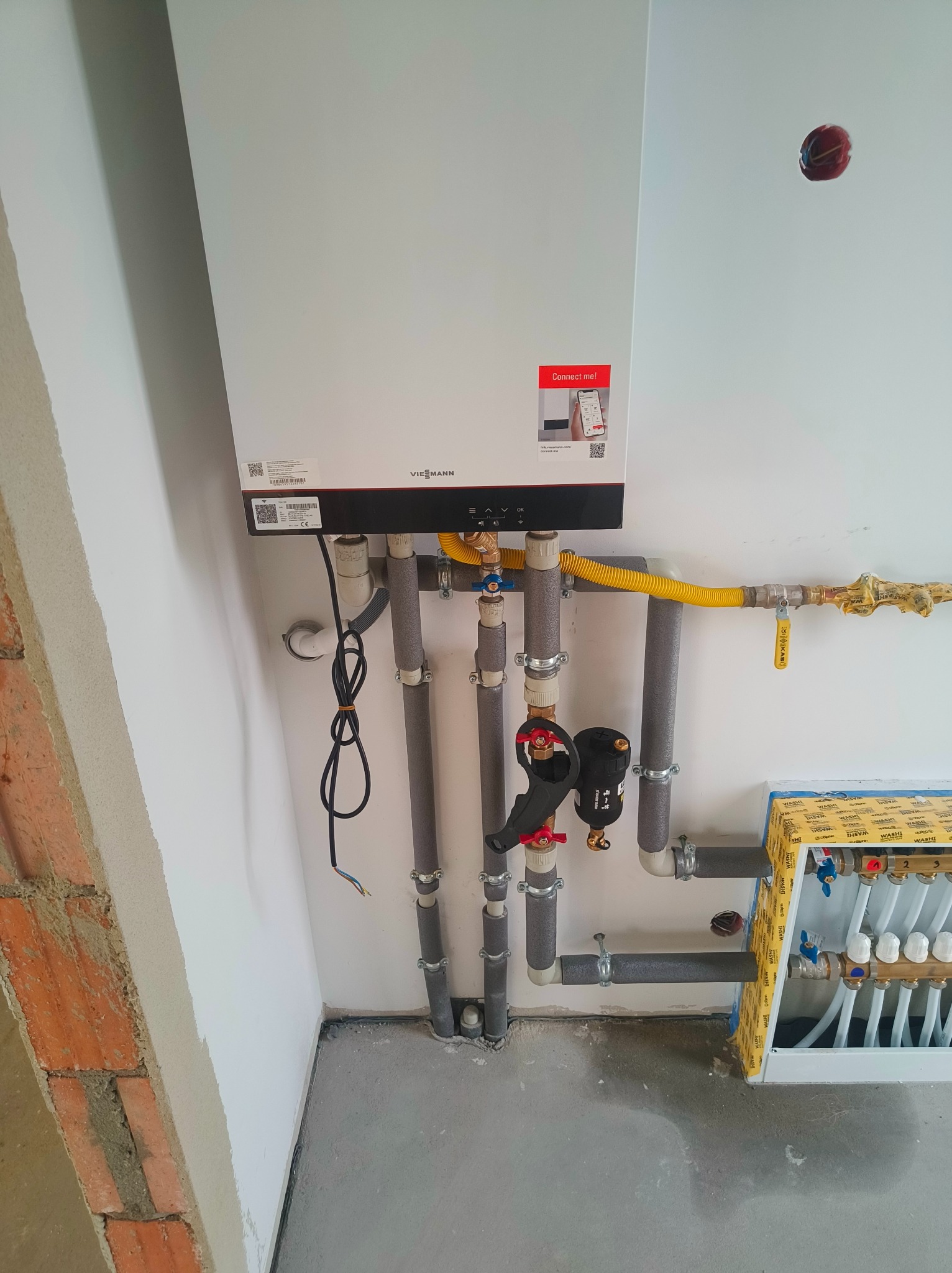 Nowoczesny piec gazowy Viessmann na białej ścianie, z widocznymi rurami instalacji grzewczej i rozdzielaczem ogrzewania podłogowego. Żółty przewód gazowy.