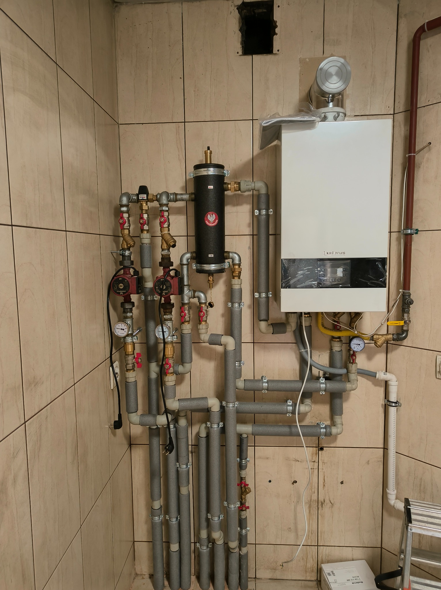 Nowoczesny piec gazowy Buderus z rozbudowaną instalacją hydrauliczną, rury zaizolowane szarą pianką, na tle beżowych płytek. Widoczne zawory i manometry.