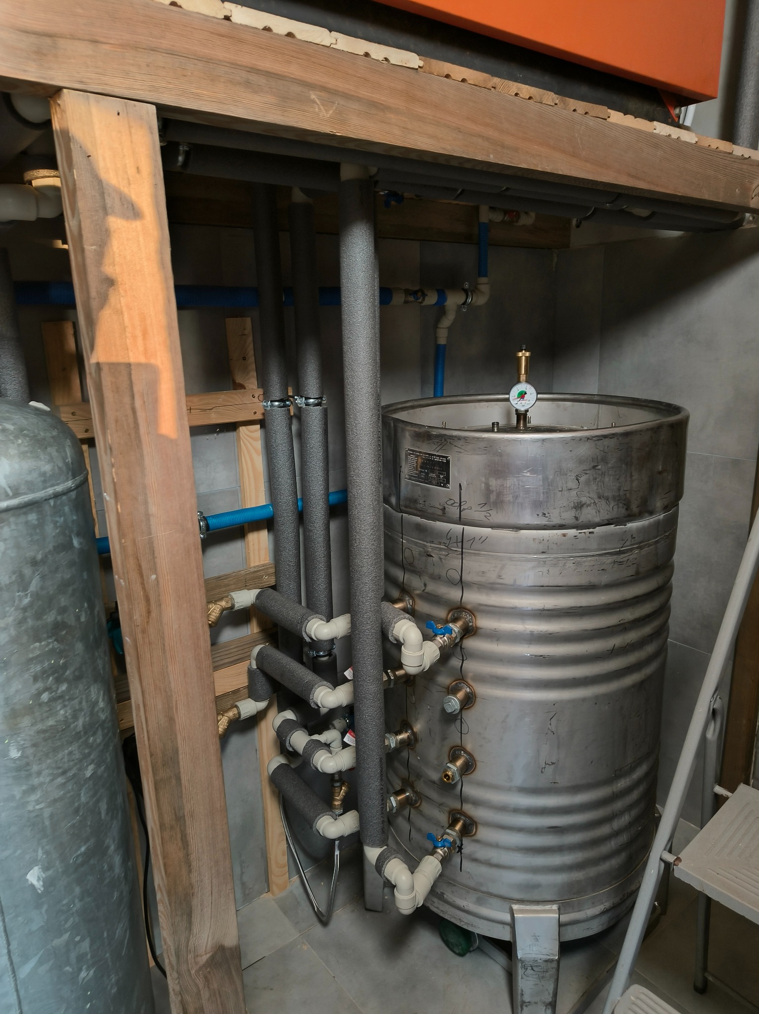 Stalowy akumulator ciepła z izolowanymi rurami w kotłowni. Widoczne liczne przyłącza i manometr. Elementy instalacji grzewczej w pomieszczeniu z szarą glazurą.