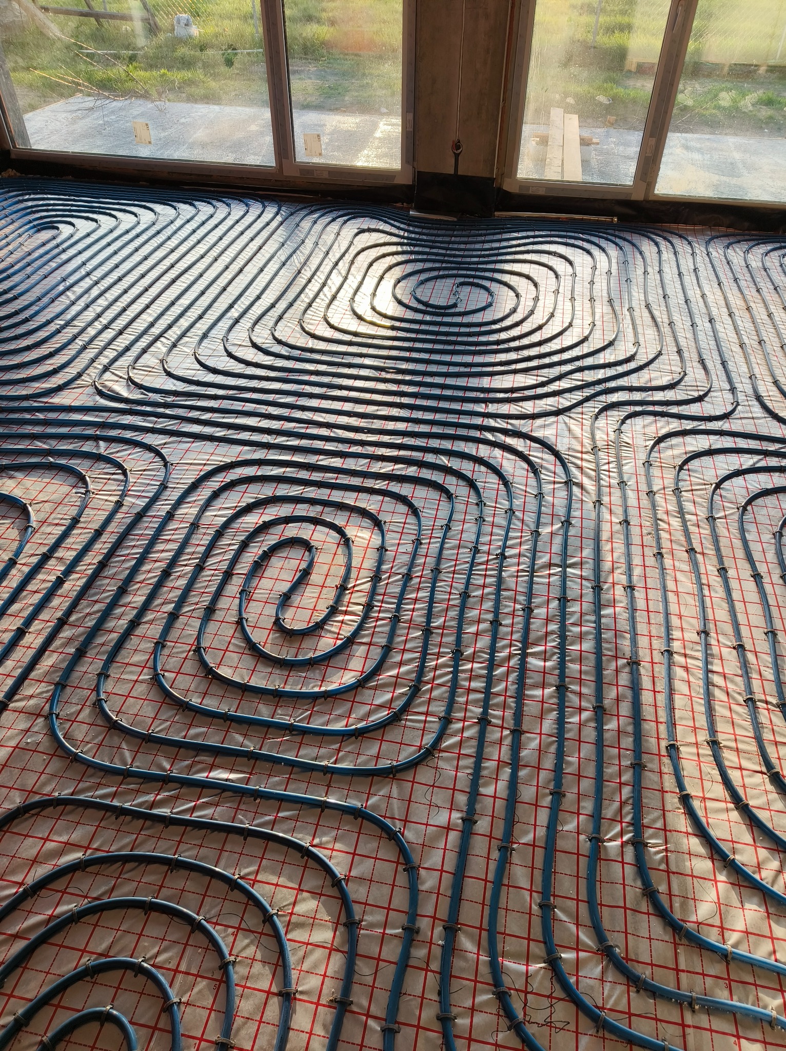 Niebieskie rury ogrzewania podłogowego ułożone spiralnie na folii z czerwoną siatką, widoczne okno i fragment surowego betonu. Oświetlenie naturalne, ciepłe.