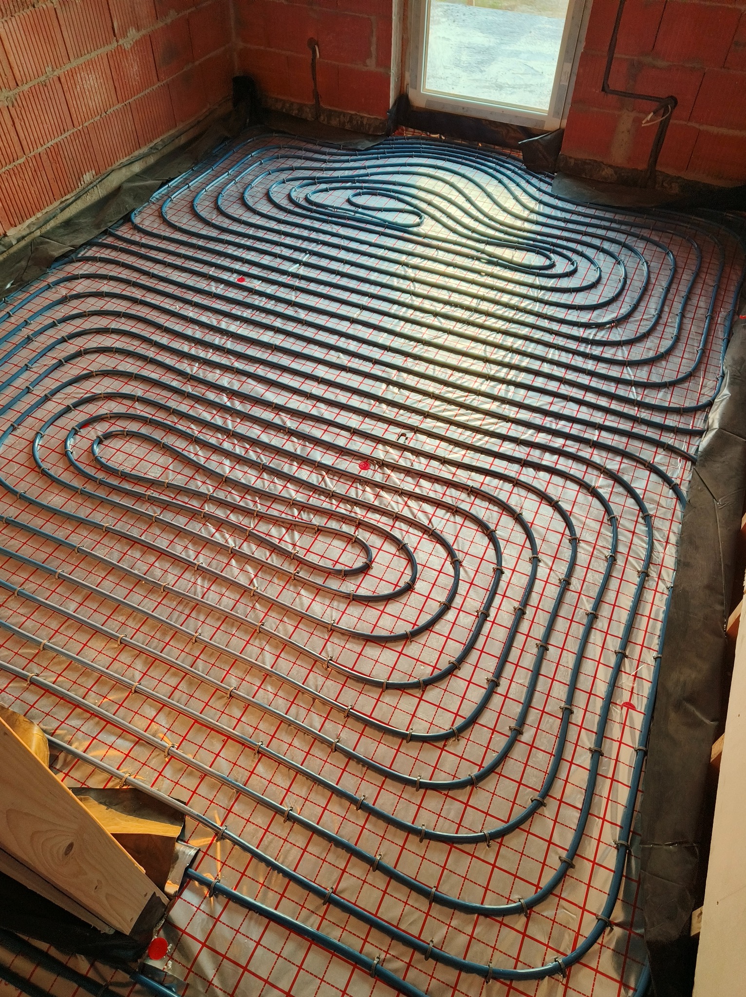 Niebieskie rury ogrzewania podłogowego ułożone spiralnie na folii z czerwoną siatką w surowym wnętrzu. Widoczne fragmenty ceglanej ściany i okno.