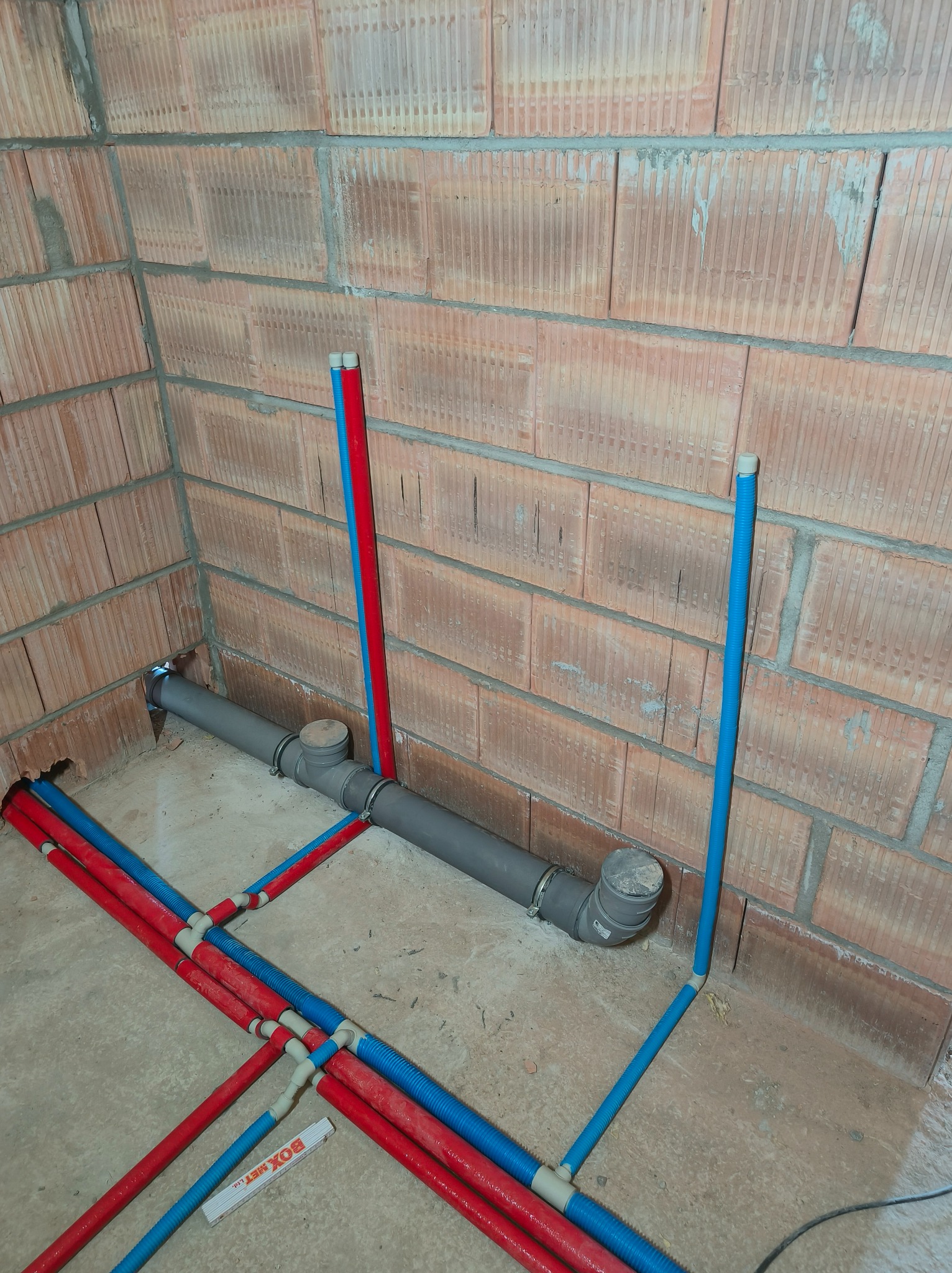 Instalacja wodna w surowym wnętrzu: rury PEX (czerwone i niebieskie) prowadzone wzdłuż ceglanej ściany i szare rury kanalizacyjne. Widoczny fragment miarki.