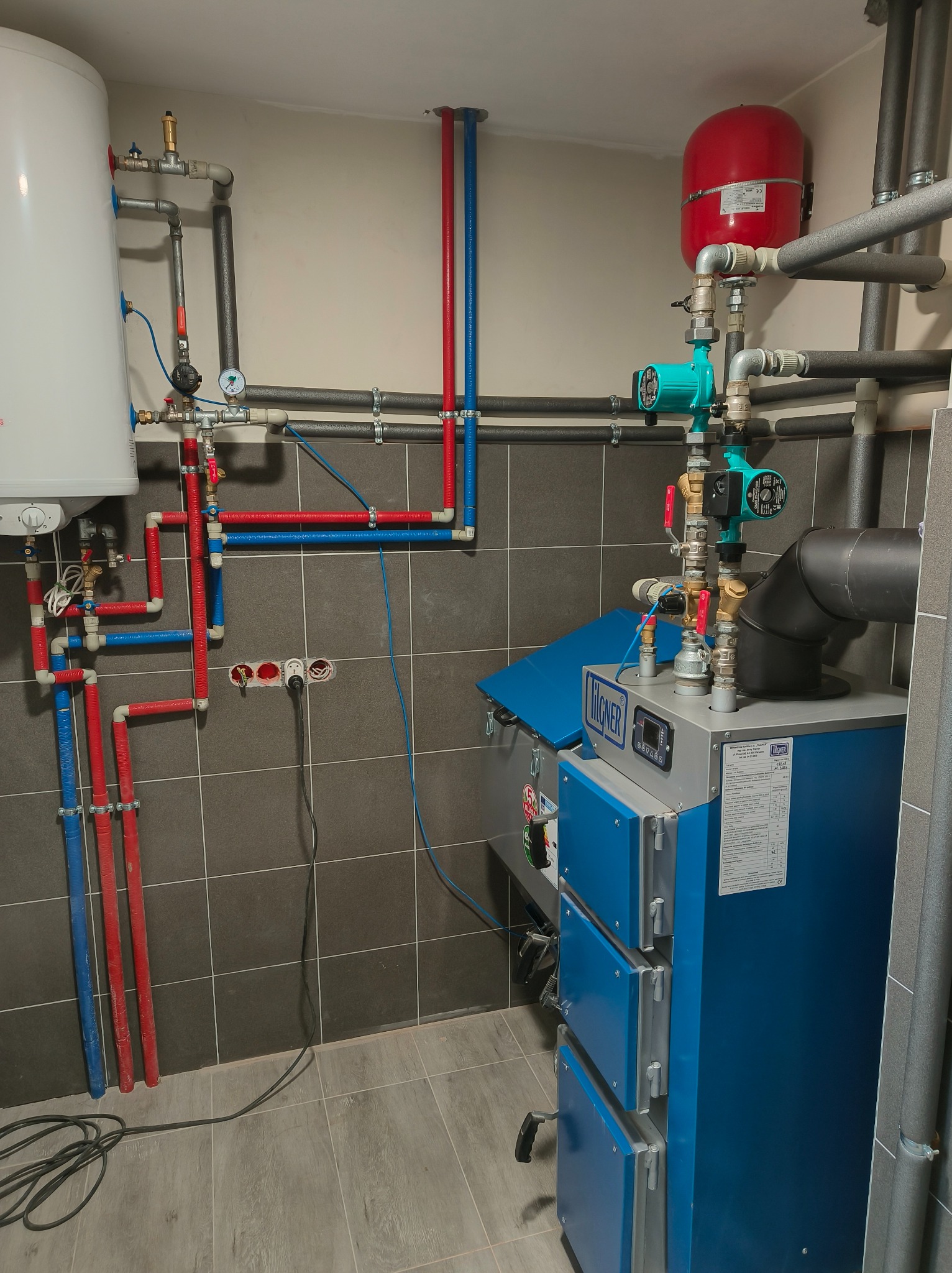 Zmontowana instalacja grzewcza z piecem Wägner, zbiornikiem, pompami obiegowymi i rurami w kolorach czerwonym i niebieskim na tle szarej ściany z płytek.