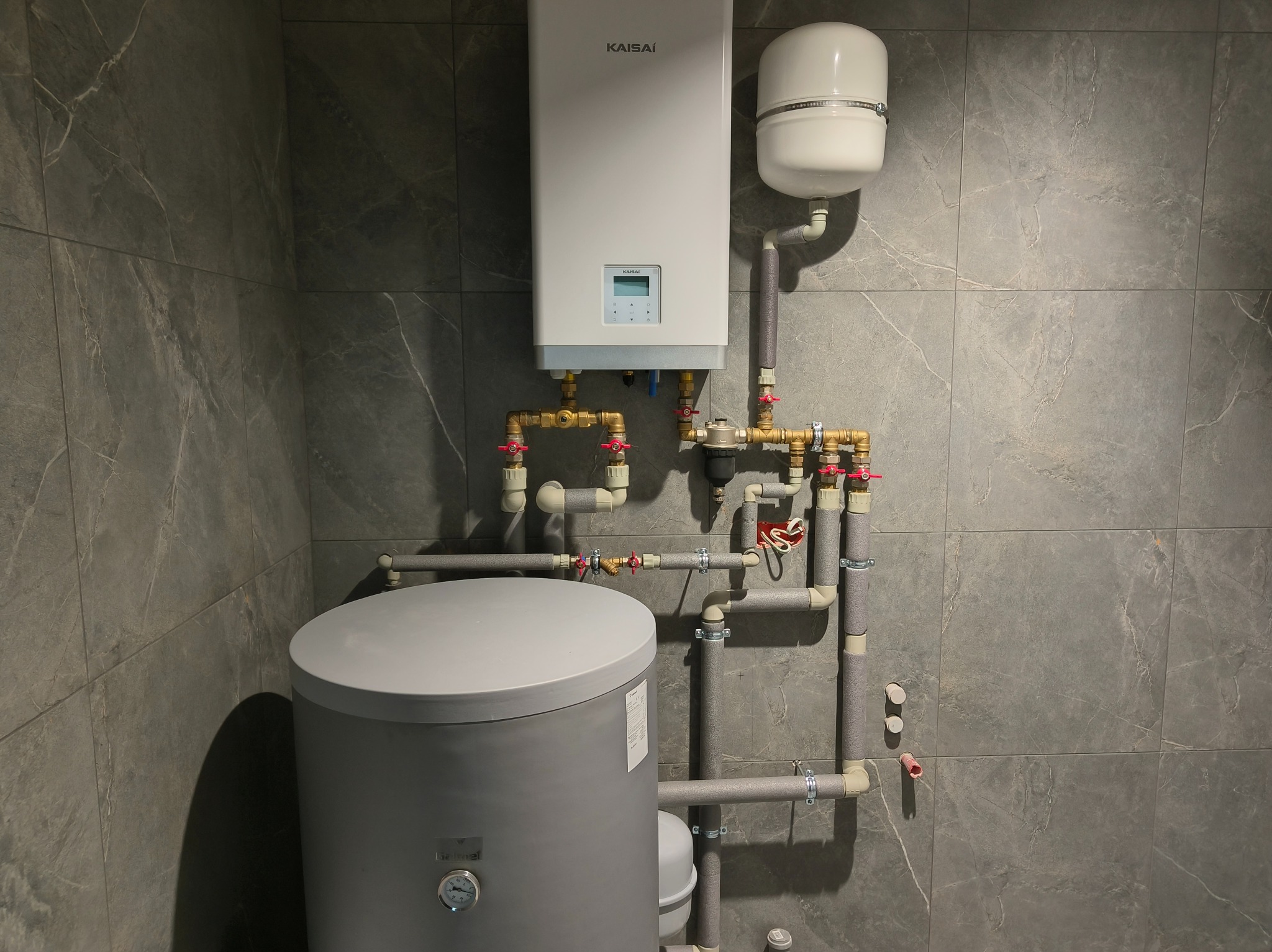 Nowoczesny piec CO marki Kaisai z szarym zasobnikiem wody, zamontowany na tle płytek imitujących marmur. Widoczne rury i zawory.