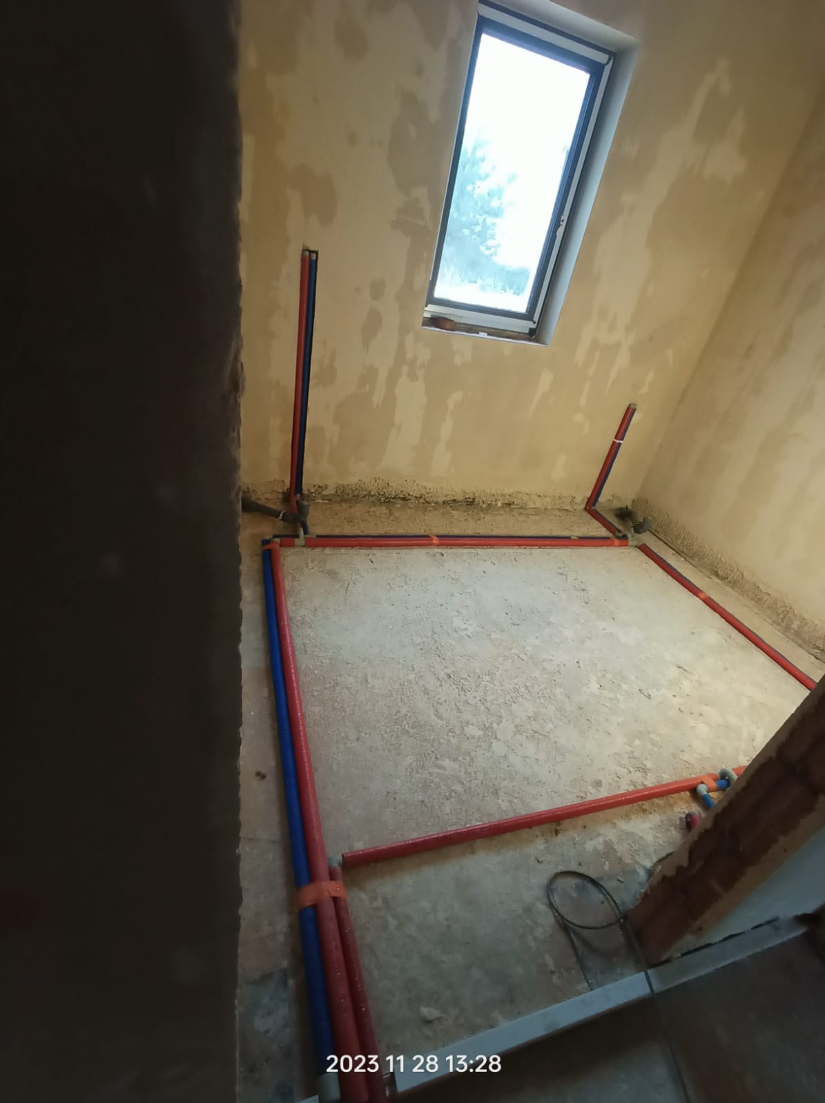 Wnętrze w trakcie prac hydraulicznych: czerwono-niebieskie rury ułożone wzdłuż ścian i pionowo, surowy beton, okno dachowe. Widok z góry, etap instalacji.