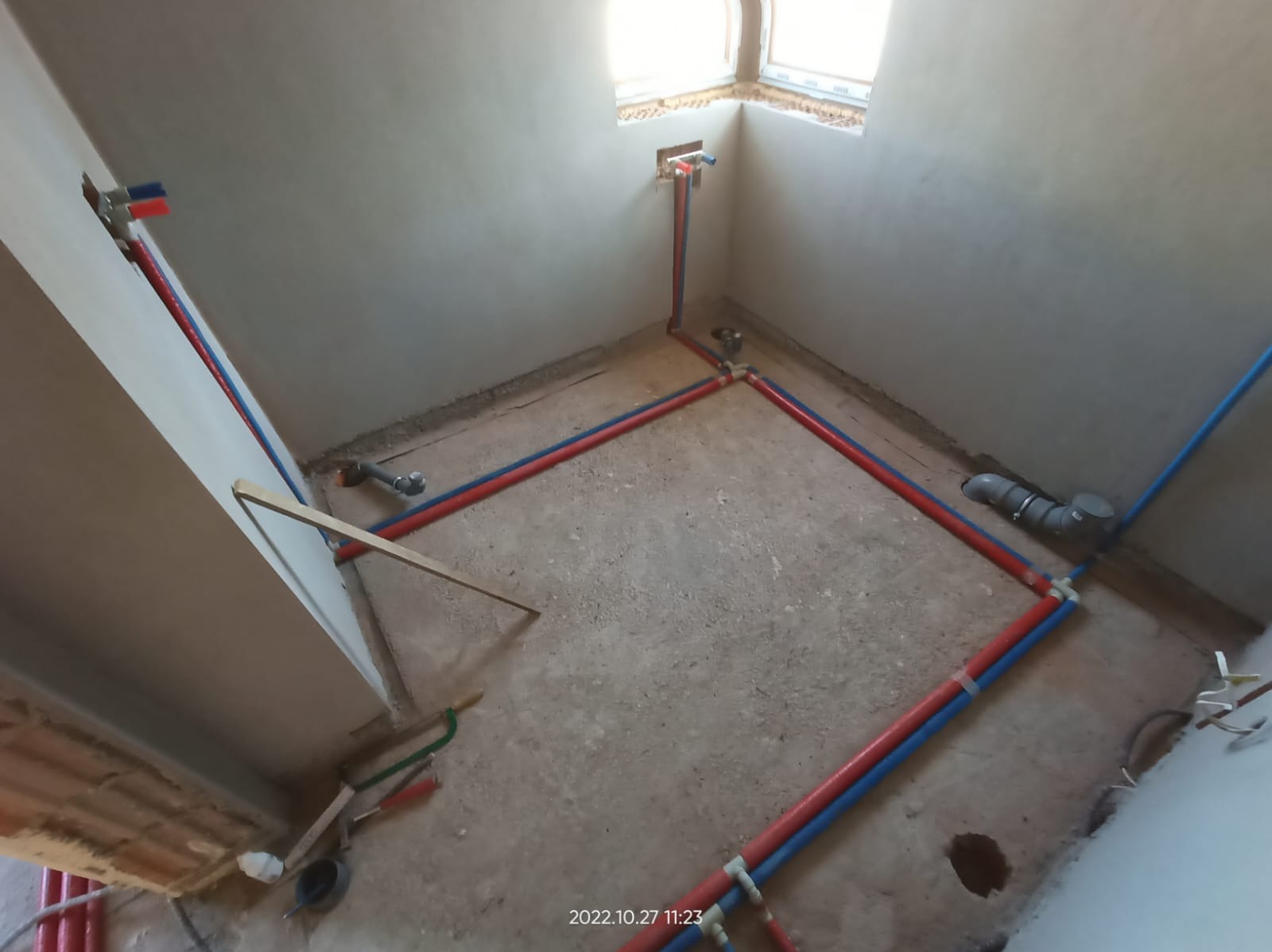 Instalacja wodna i kanalizacyjna w surowym pomieszczeniu. Rury PEX w kolorze niebieskim i czerwonym ułożone wzdłuż ścian i pod oknem. Widoczne narzędzia hydrauliczne.