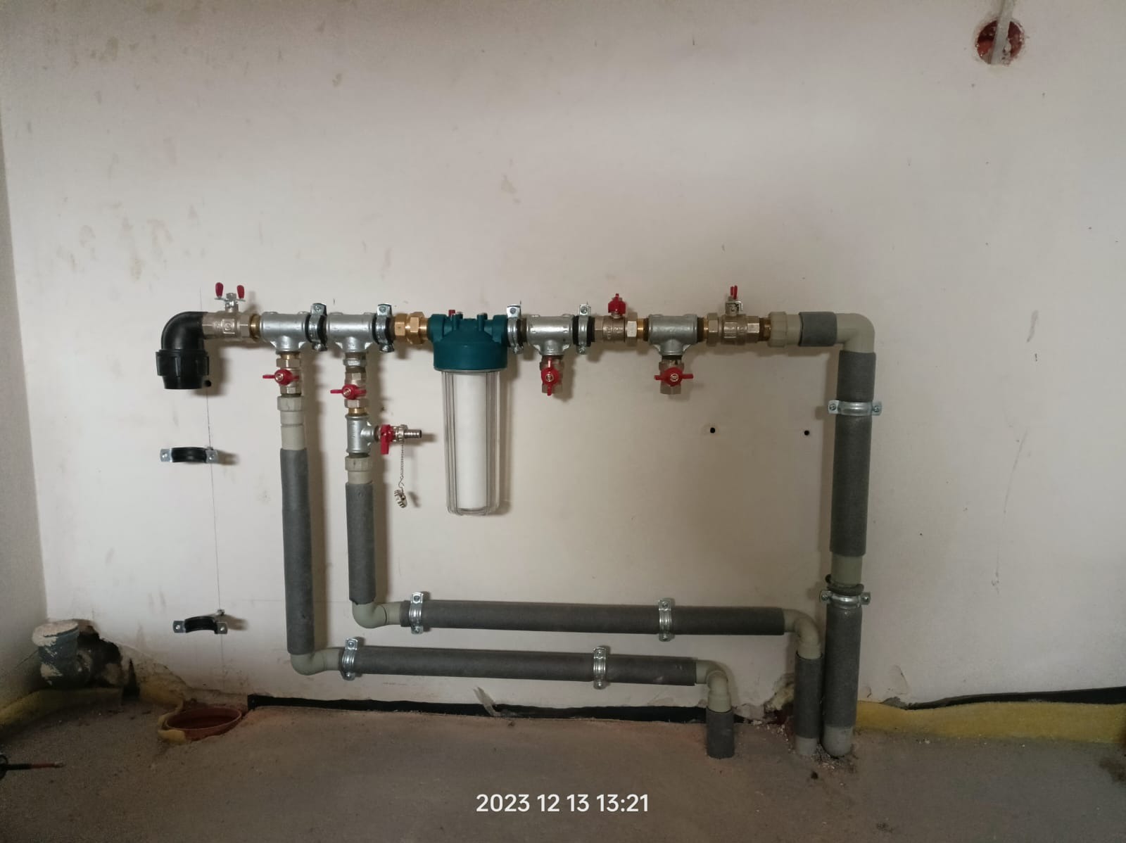 Szara instalacja wodna z filtrem i zaworami na białej ścianie. Rury mocowane obejmami. Widoczne przyłącza i elementy instalacji. Surowy stan wykończenia wnętrza.