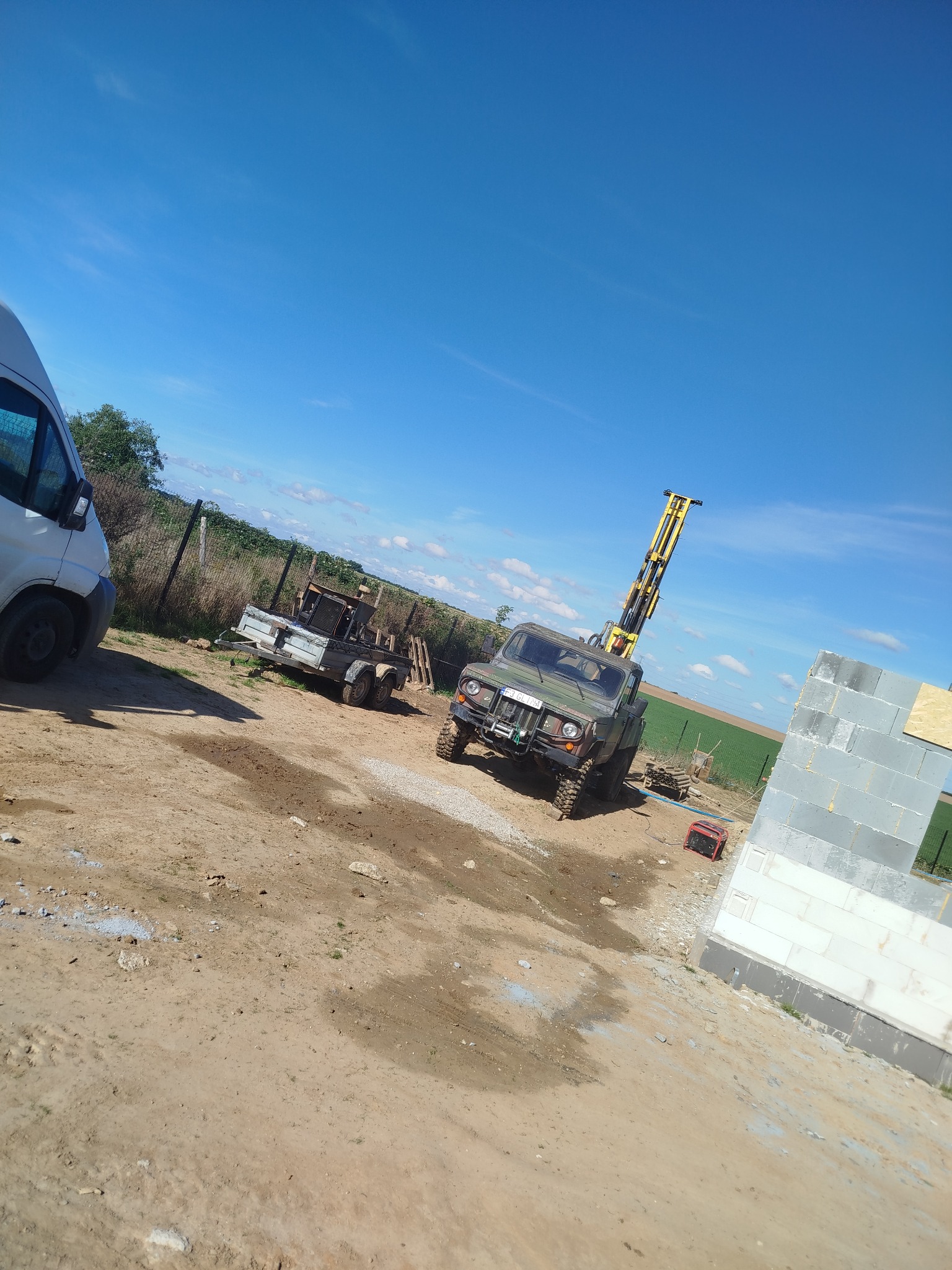 Wiertnica na podwoziu samochodu terenowego na placu budowy, obok przyczepa i fragment ściany z bloczków. Błękitne niebo w tle. Wiertnica gotowa do pracy.