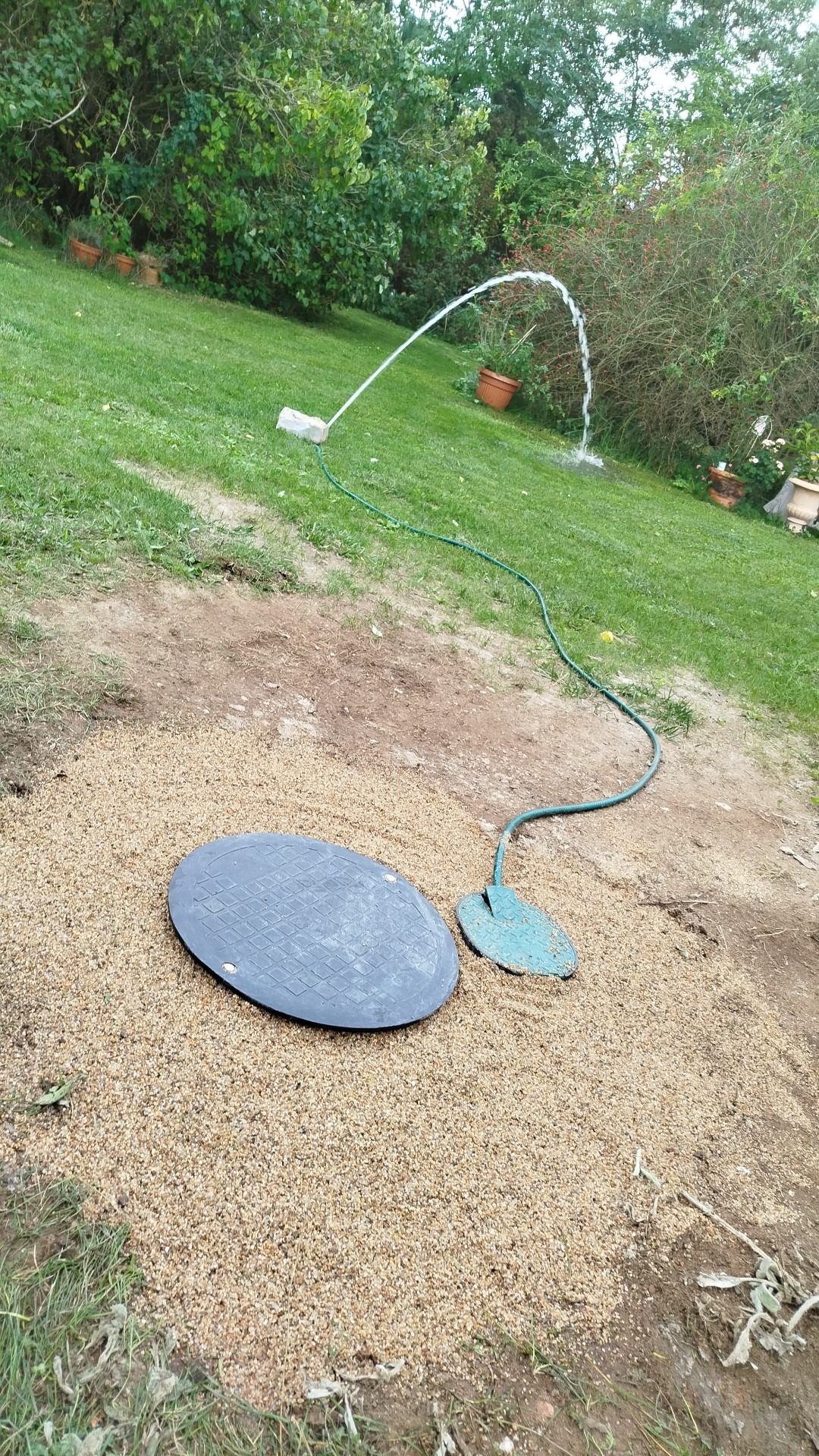 Studnia z pokrywą, zasilana wężem ogrodowym, tworząca fontannę na trawniku. Widoczne rośliny w tle, ziemia wokół studni wysypana drobnym żwirem.