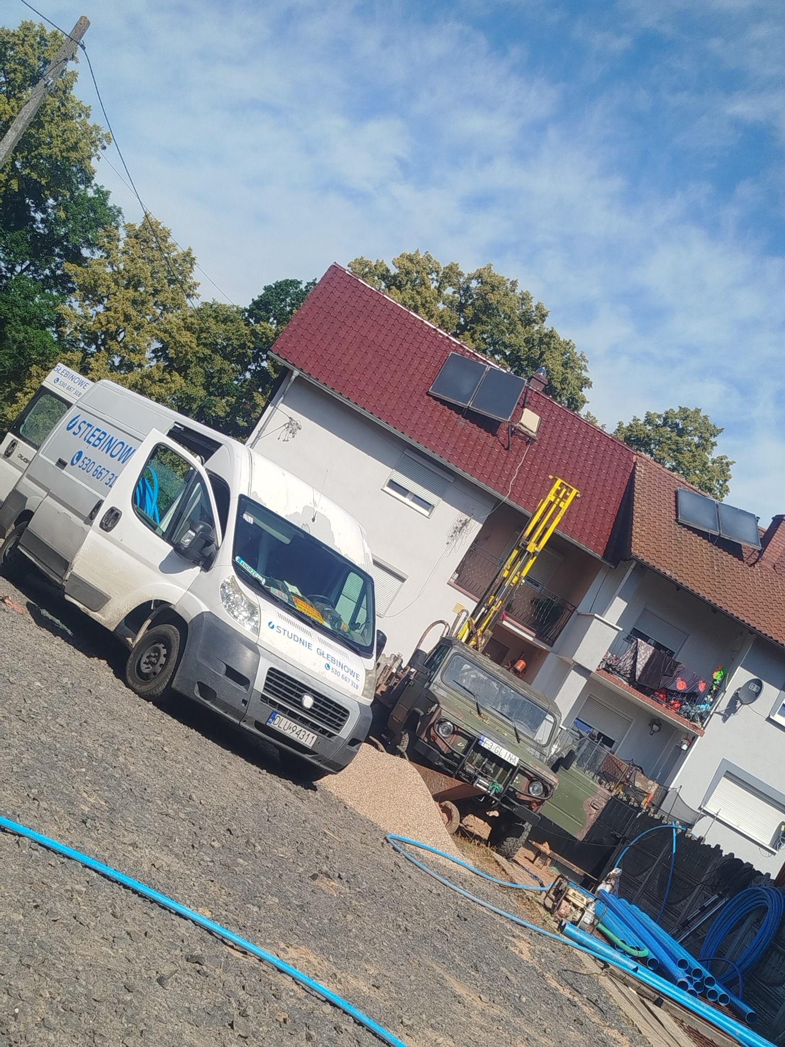 Realizacja studni głębinowej: biały bus z logo firmy 'Studnie Głębinowe' i sprzęt wiertniczy na tle domu z panelami słonecznymi i błękitne rury.