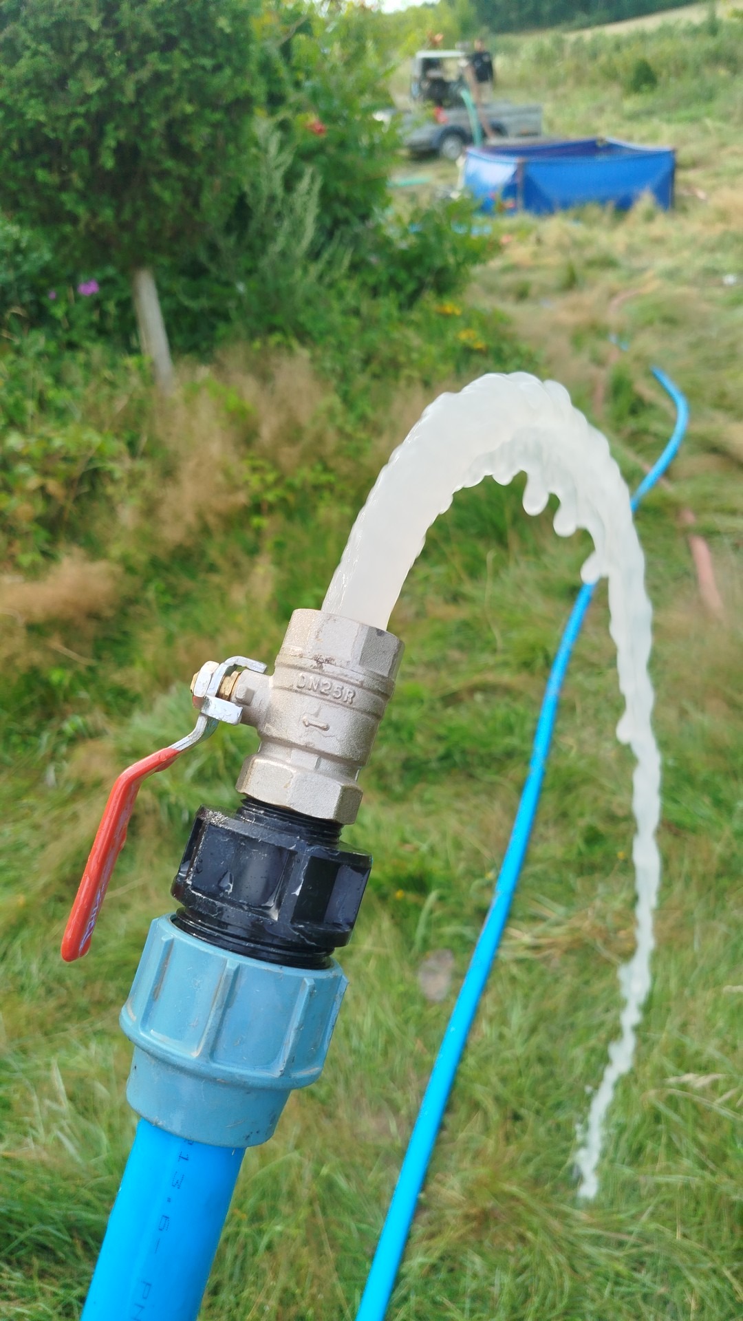 Woda wypływająca z niebieskiego węża podłączonego do zaworu DN25R, na tle zielonej trawy i roślinności. W tle widoczny zbiornik na wodę i sprzęt.