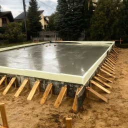 Marcin Kwaśny - Zalany fundament z izolacją i drewnianym szalunkiem, widok z góry. Świeży beton odbija światło, w tle domy i drzewa. Etap budowy domu.