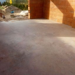 Marcin Kwaśny - Surowy beton wewnątrz budynku z cegły ceramicznej, widoczny piasek i taczka na zewnątrz. Świeżo wylana posadzka w trakcie budowy domu jednorodzinnego.