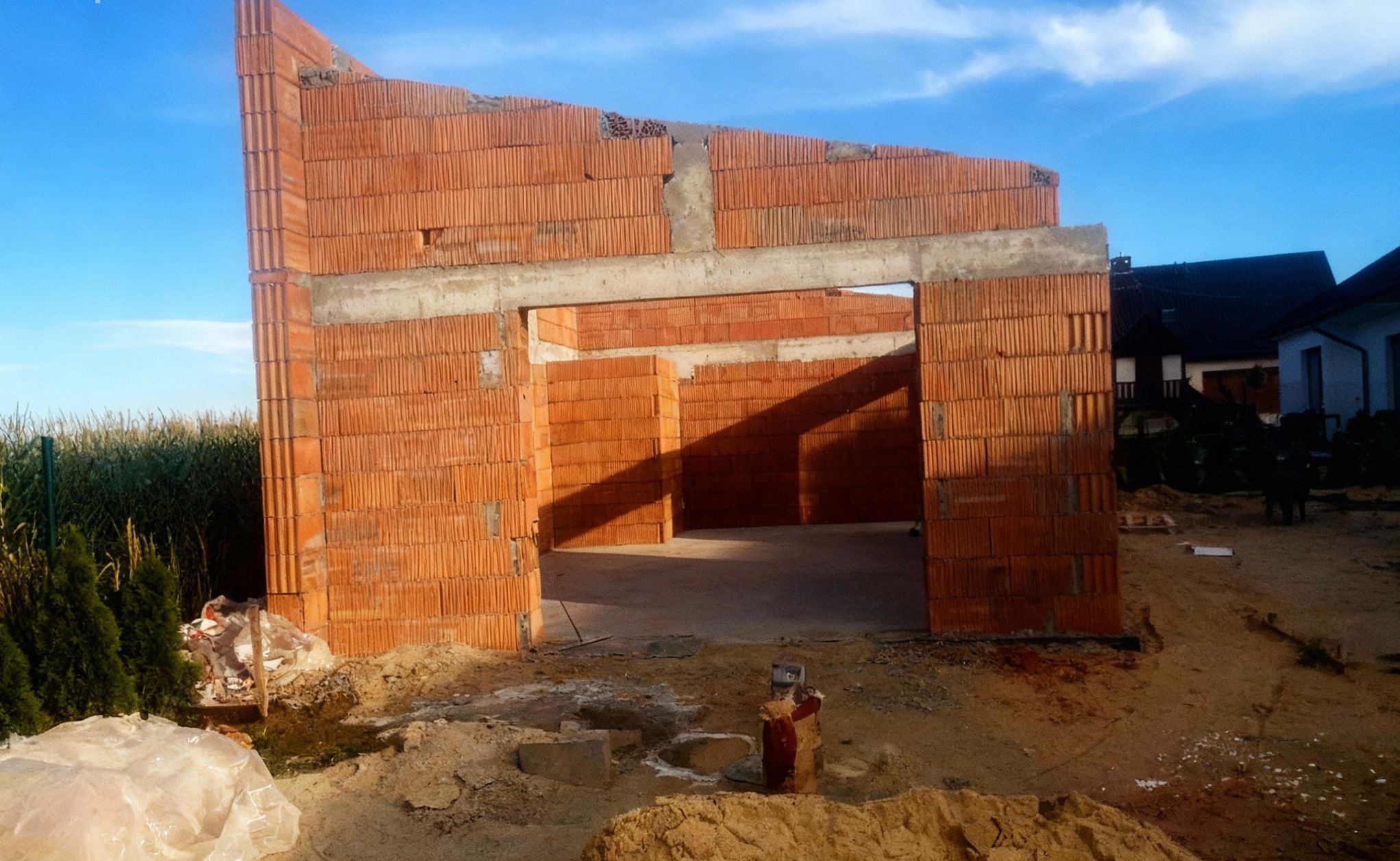 Wznoszenie ścian z czerwonej cegły na budowie domu jednorodzinnego. Widoczne zbrojenia i betonowe nadproża. W tle zabudowa jednorodzinna i błękitne niebo.