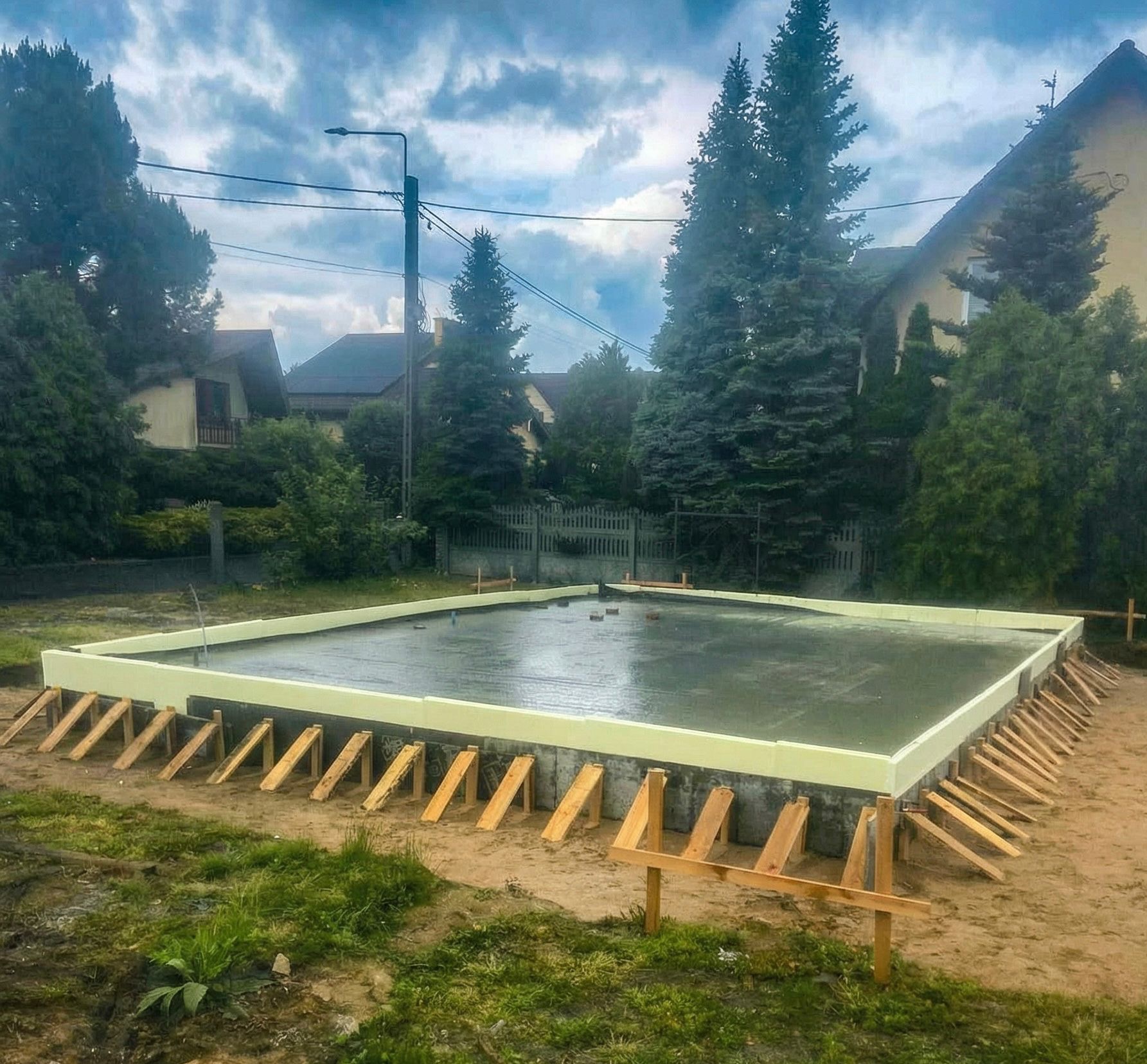 Zalany fundament z izolacją i drewnianym szalunkiem na tle zieleni i zabudowań. Widok z poziomu gruntu, pochmurne niebo.