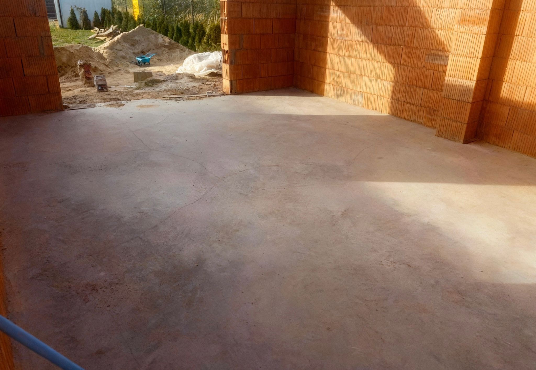 Surowy beton wewnątrz budynku z cegły ceramicznej, widoczny piasek i taczka na zewnątrz. Świeżo wylana posadzka w trakcie budowy domu jednorodzinnego.