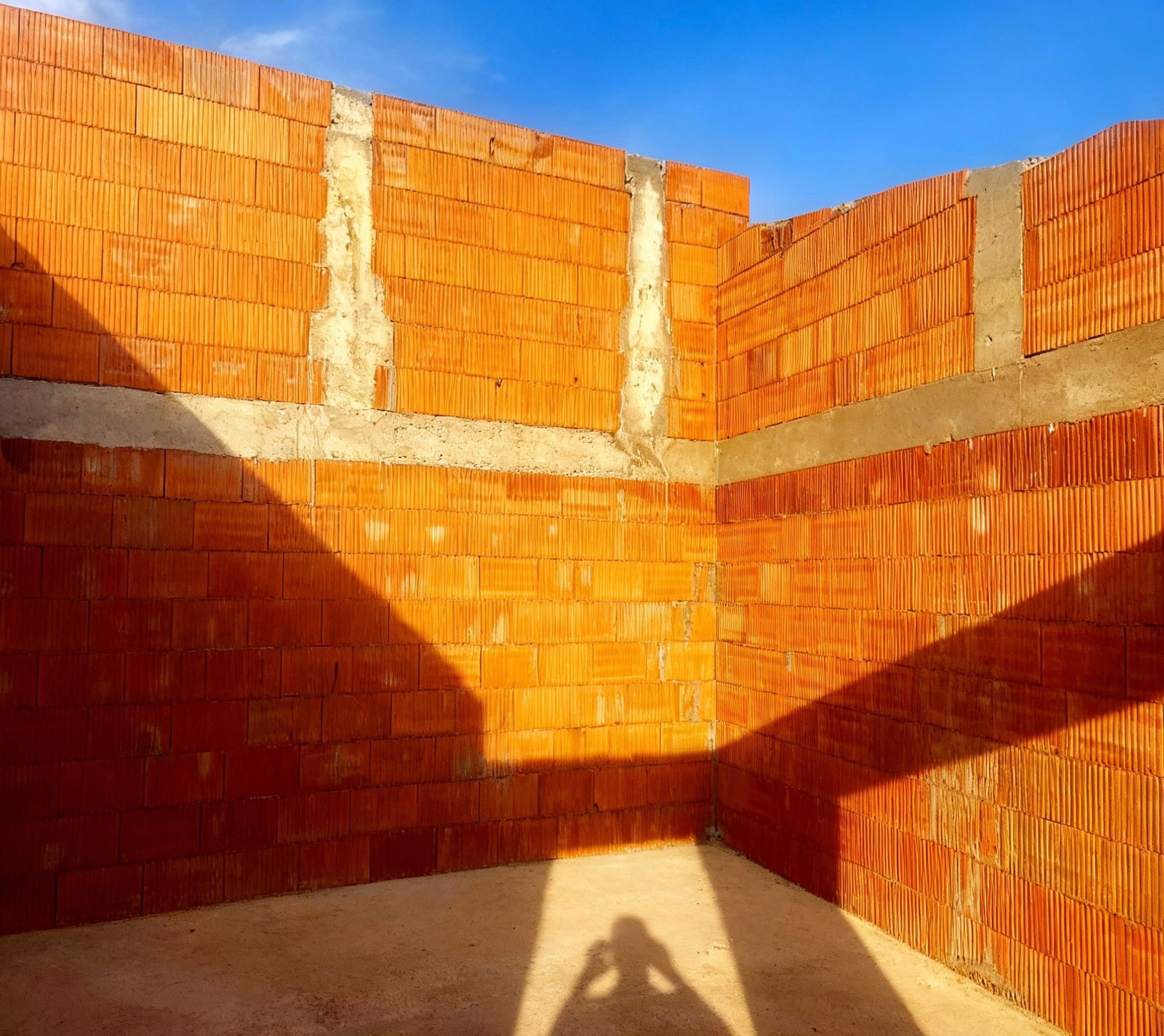 Wnętrze budynku w trakcie budowy: ściany z czerwonej cegły ceramicznej, widoczne spoiny i cień osoby fotografującej na podłodze. Słoneczny dzień, błękitne niebo w tle.