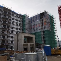 jbngroup - Budynek w trakcie budowy, z rusztowaniami, siatkami ochronnymi i banerami reklamowymi, widoczne materiały budowlane i przenośna toaleta.