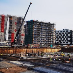 jbngroup - Budowa wielopiętrowego budynku mieszkalnego z rusztowaniami i dźwigiem, widoczni pracownicy w odblaskowych kamizelkach, zbrojenie fundamentów na pierwszym planie.